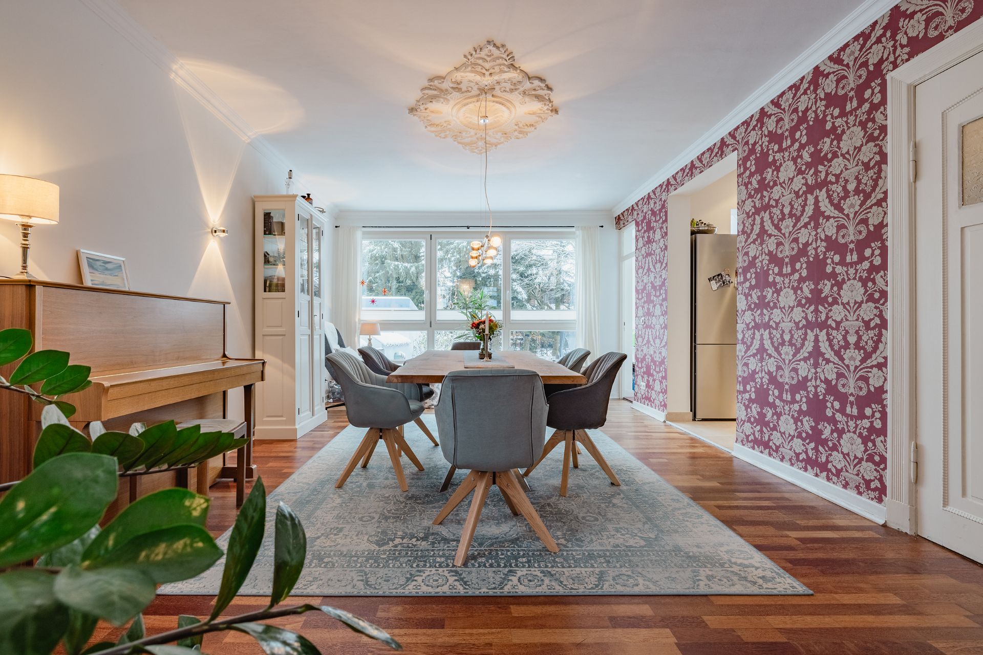 Esszimmer mit Holztisch, sechs Stühlen, Klavier, großer Fensterfront und gemusterter Tapete.