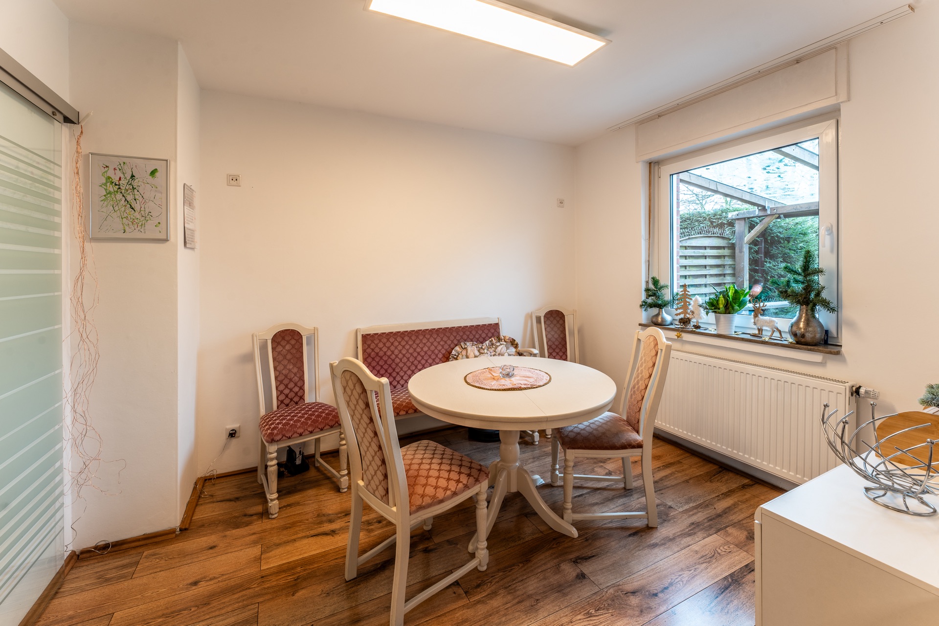 Essbereich mit rundem Tisch, vier Stühlen, Bank und Fenster mit Pflanzen auf der Fensterbank.
