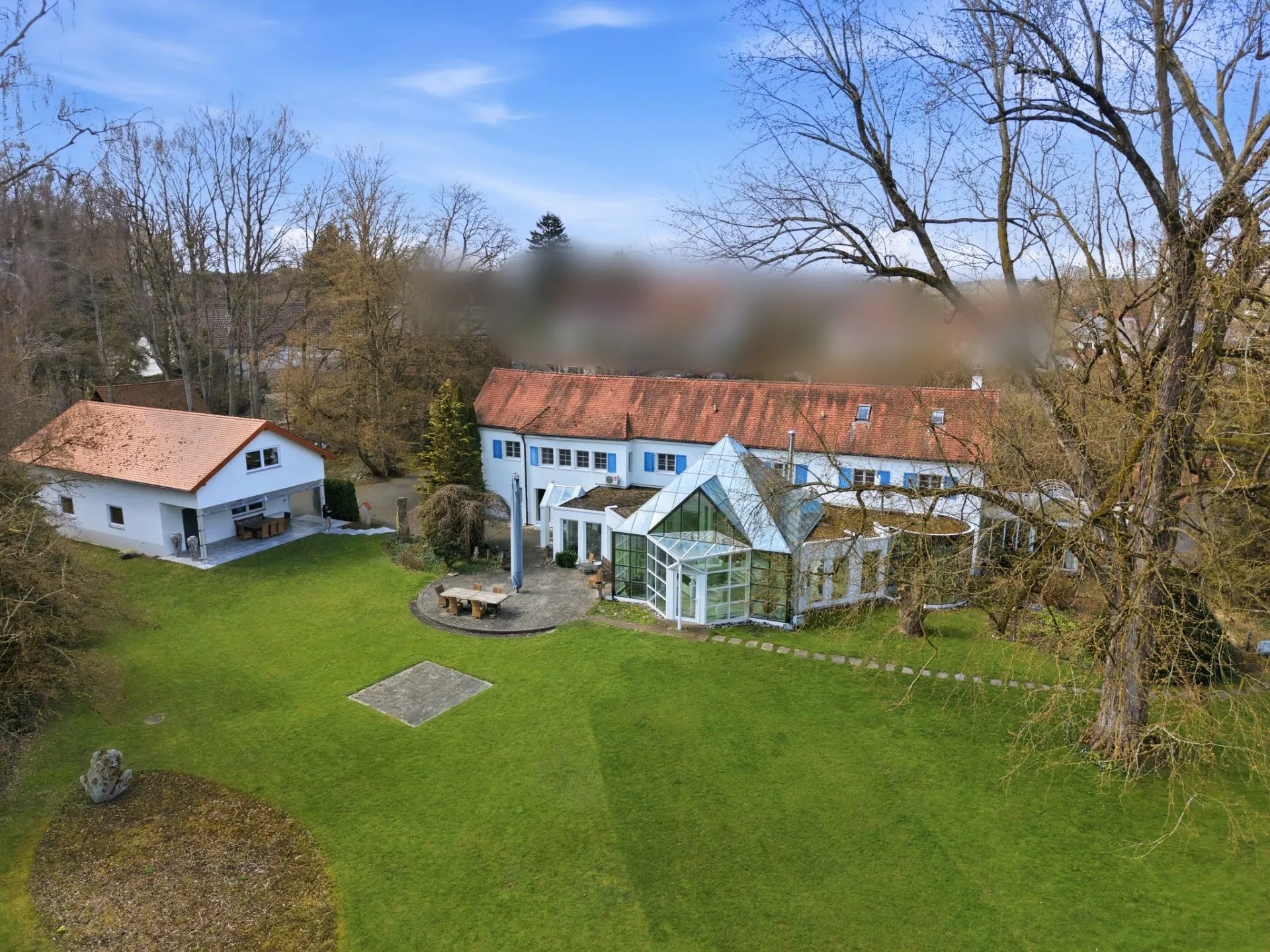 Großes Wohnhaus mit Wintergarten, Nebengebäude und großer Rasenfläche, von Bäumen umgeben.