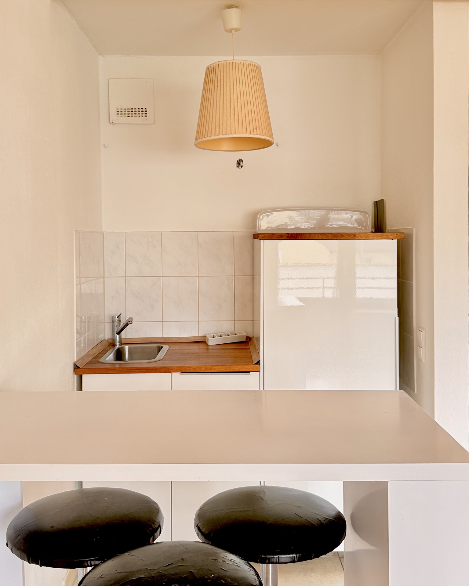 Kleine Küche mit Spüle, Kühlschrank, Theke und drei schwarzen Hockern.