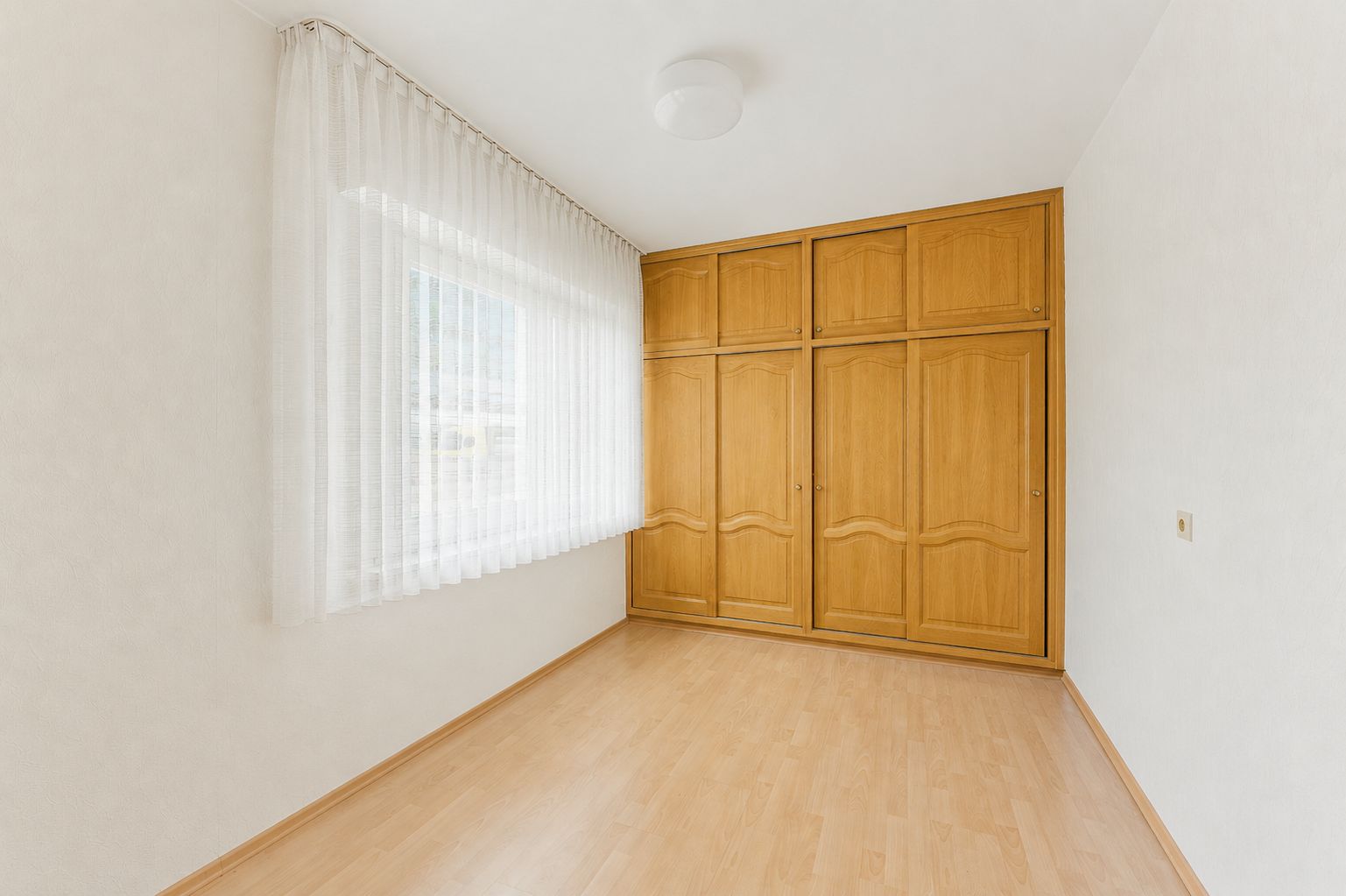 Heller Raum mit großem Fenster, hellen Vorhängen und eingebautem Holzkleiderschrank.