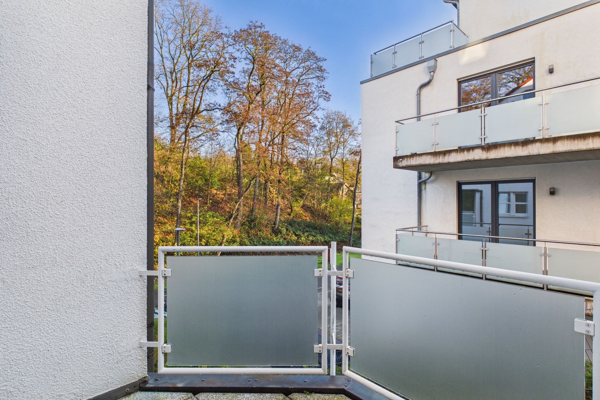 Balkon mit Milchglasgeländer, daneben modernes Wohngebäude und Bäume im Hintergrund.