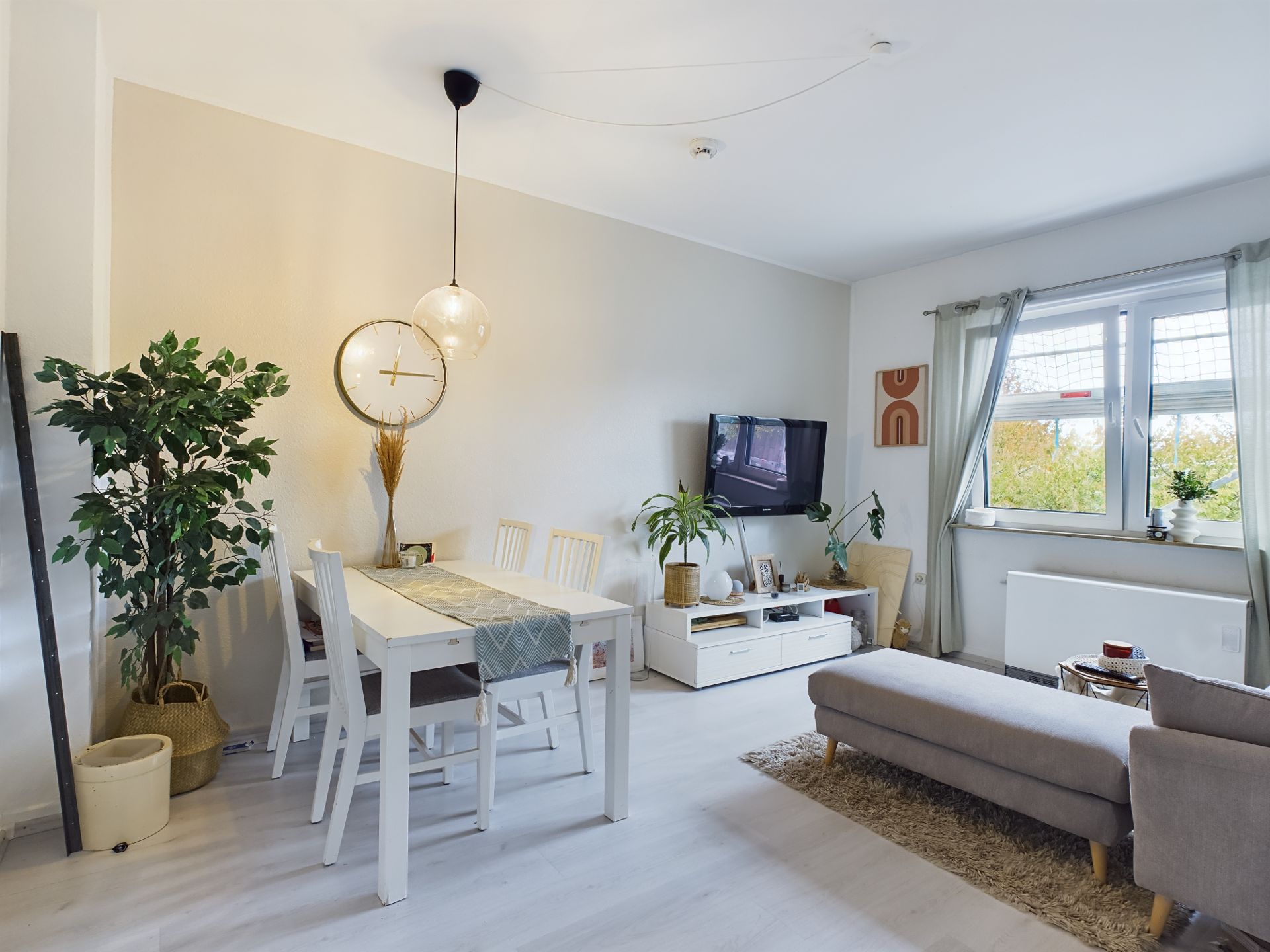Helles Wohnzimmer mit Esstisch, Fernseher, Pflanzen und großem Fenster mit Vorhängen.