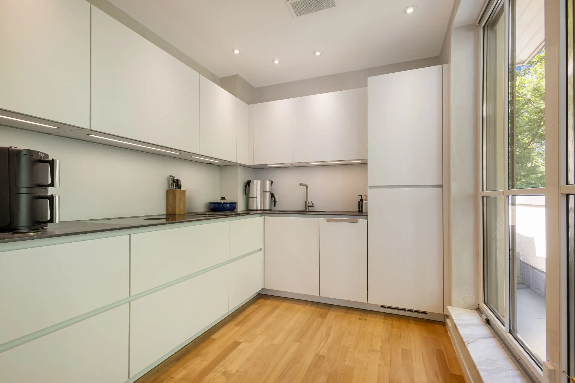 Moderne Einbauküche mit weißen Schränken, Geräten und großem Fenster, Holzboden.