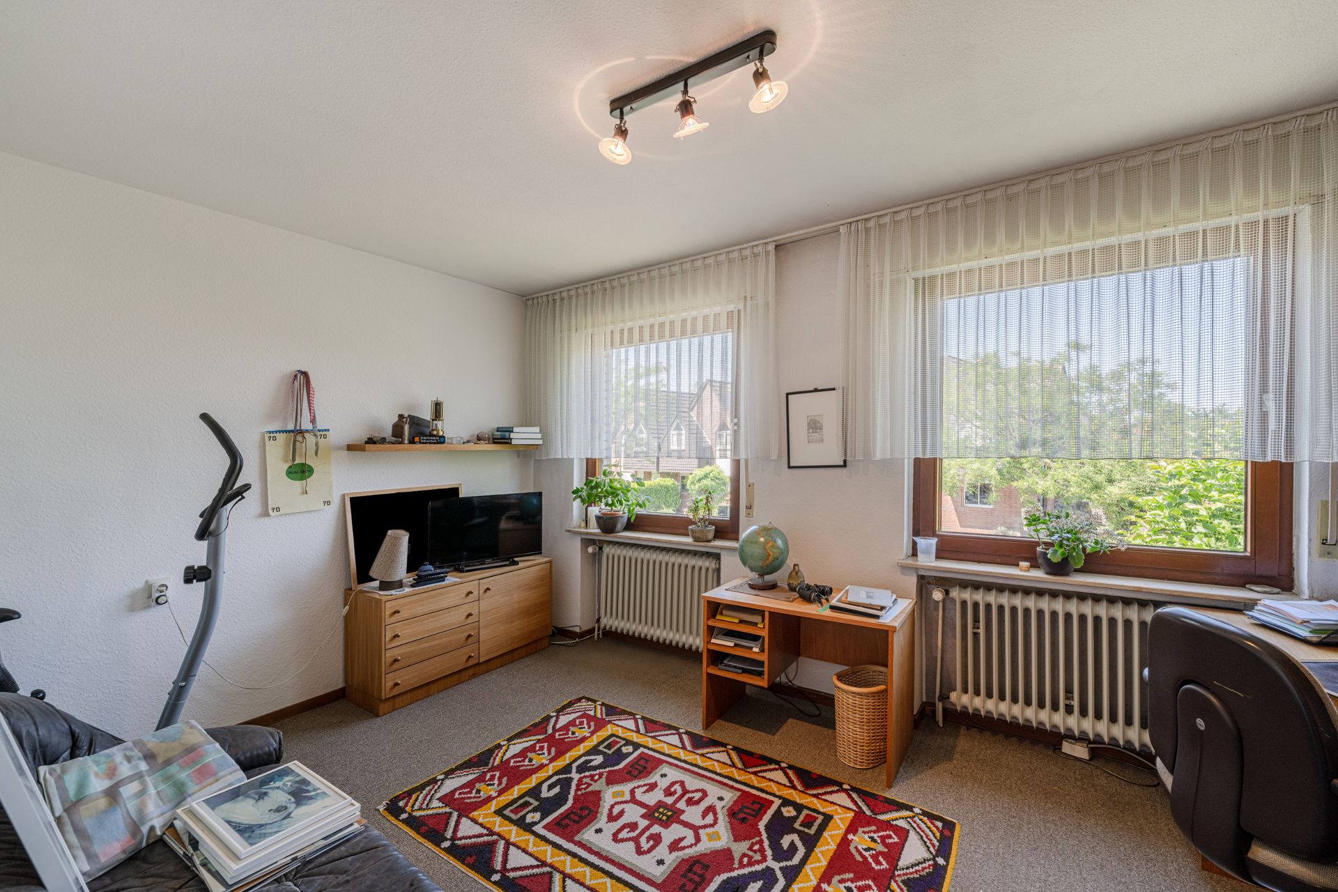 Helles Arbeitszimmer mit Schreibtisch, TV, Teppich, Pflanzen und großen Fenstern mit Gardinen.