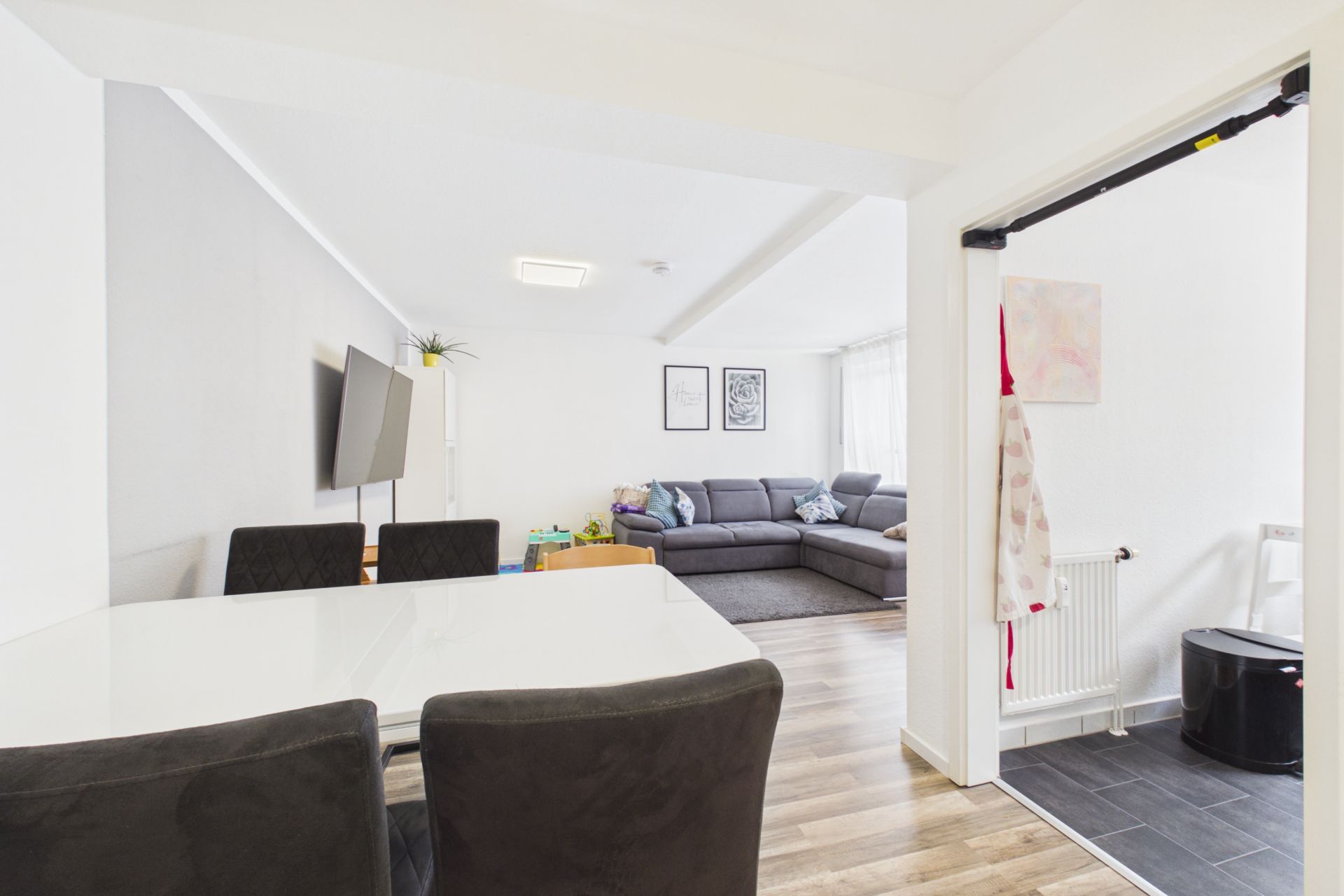 Essbereich mit weißem Tisch, Blick ins Wohnzimmer mit grauem Sofa und Fernseher.