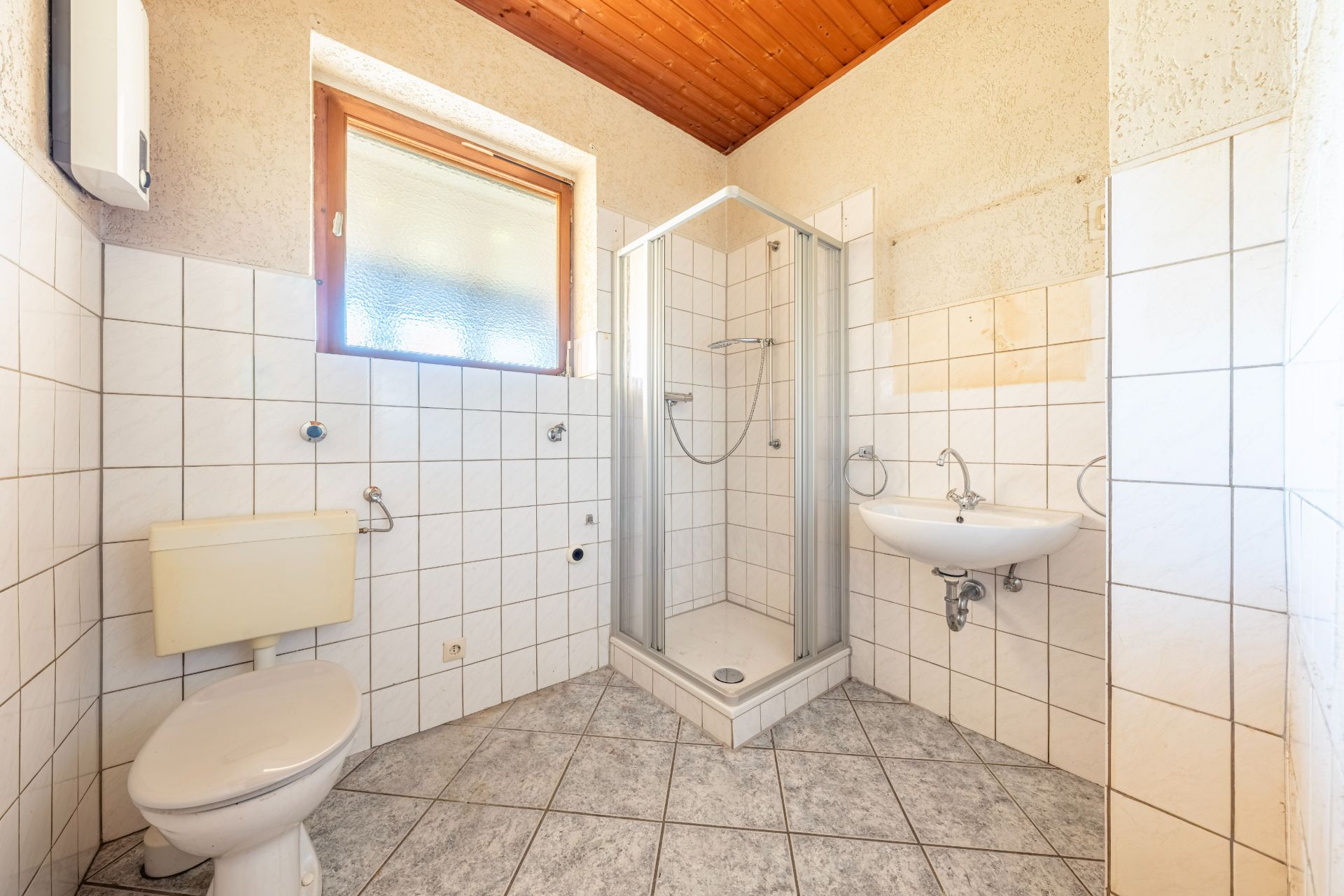 Helles Badezimmer mit Dusche, WC, Waschbecken und Fenster, gefliest und mit Holzdecke.