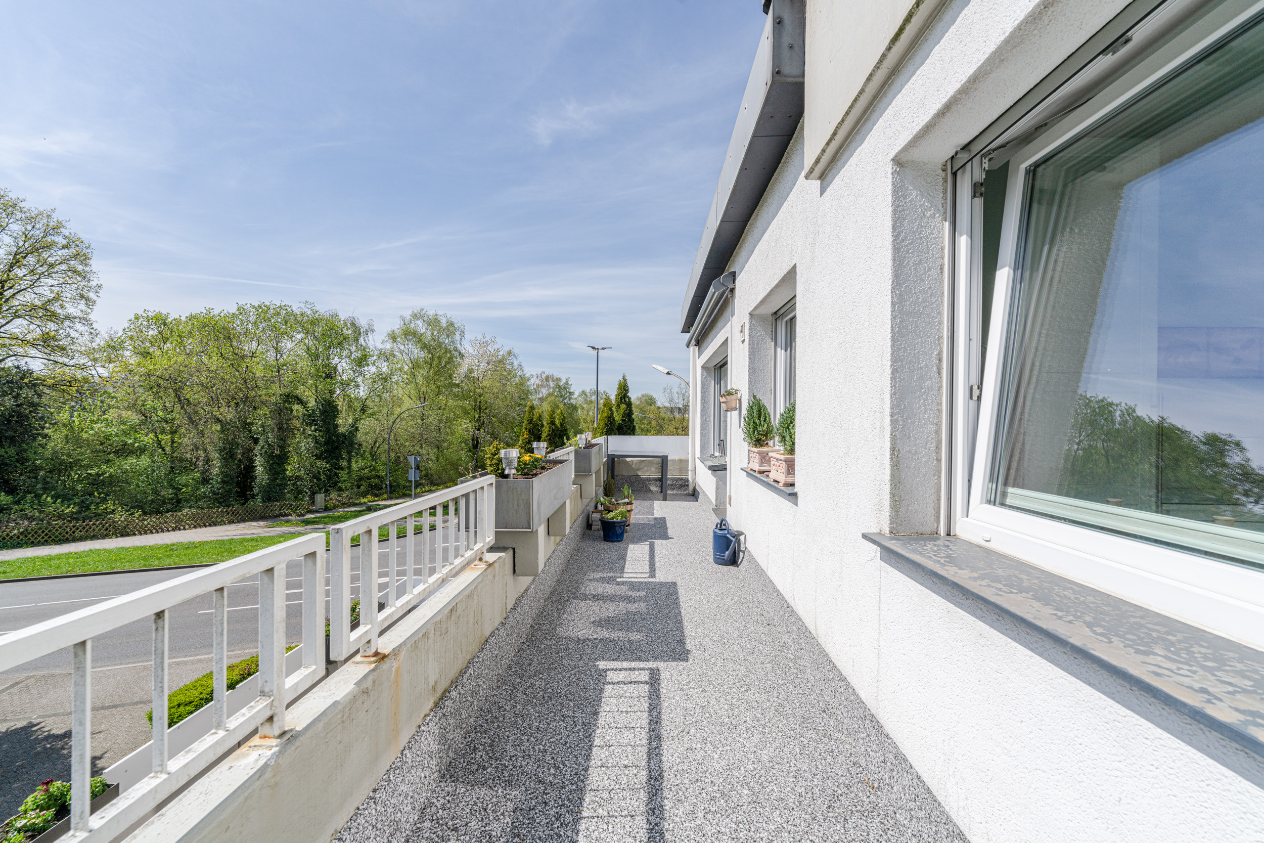 Weite Terrasse mit grauem Boden, weißen Geländern und Blumenkübeln neben einem Wohnhaus.