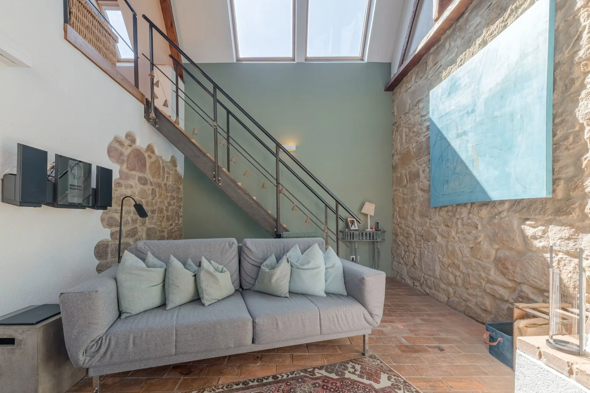 Wohnzimmer mit grauem Sofa, mehreren Kissen, gemauerter Wand und Treppe mit Metallgeländer.
