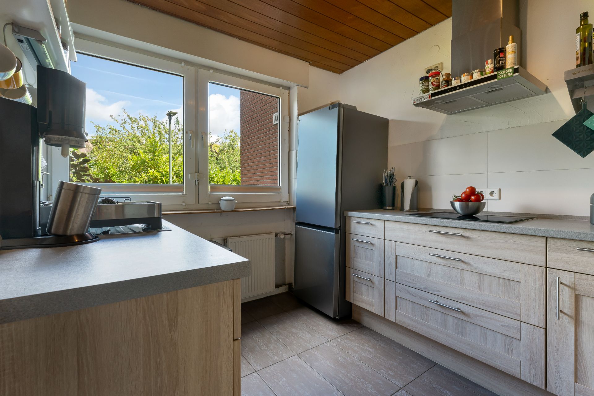 Helle Küchenzeile mit grauem Kühlschrank, Holzfronten und großen Fenstern mit Blick ins Grüne.