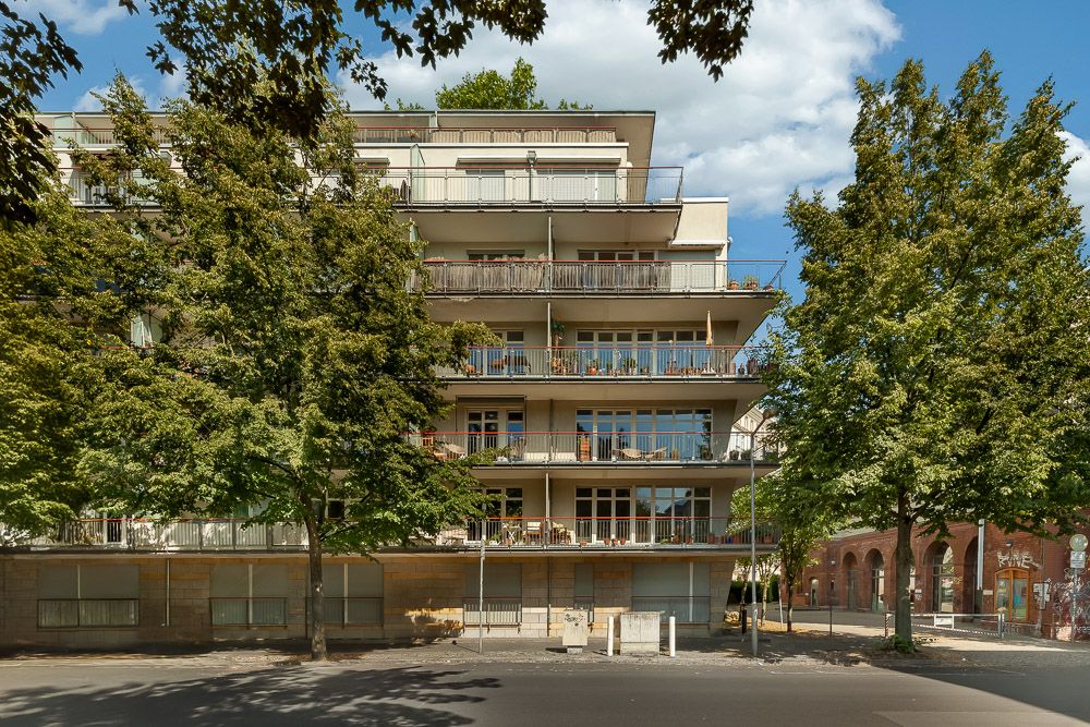 Mehrstöckiges Wohngebäude mit großen Balkonen, umgeben von Bäumen an einer Straße.
