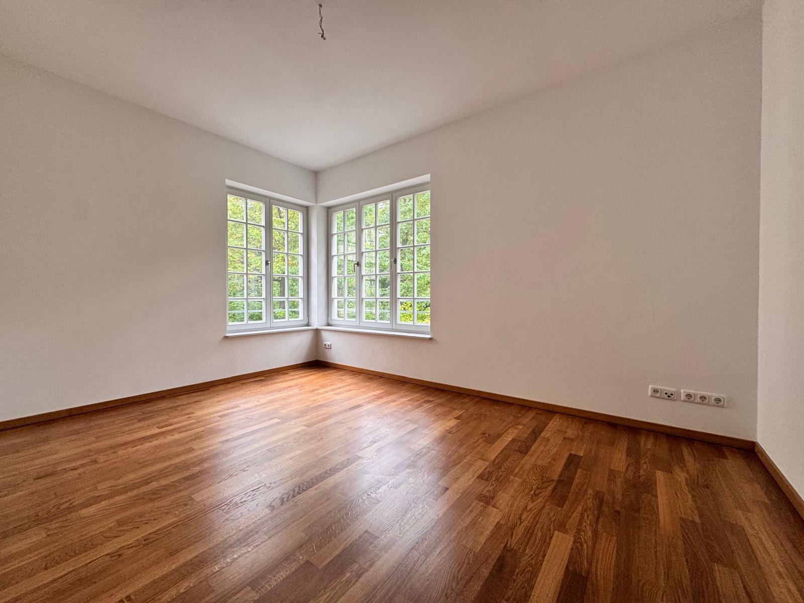 Leerer Raum mit Parkettboden und großen Eckfenstern, weiße Wände und Steckdosen an der Wand.