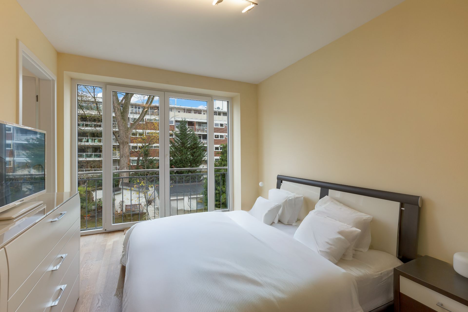 Helles Schlafzimmer mit Doppelbett, großem Fenster und Blick auf Bäume und Wohngebäude.