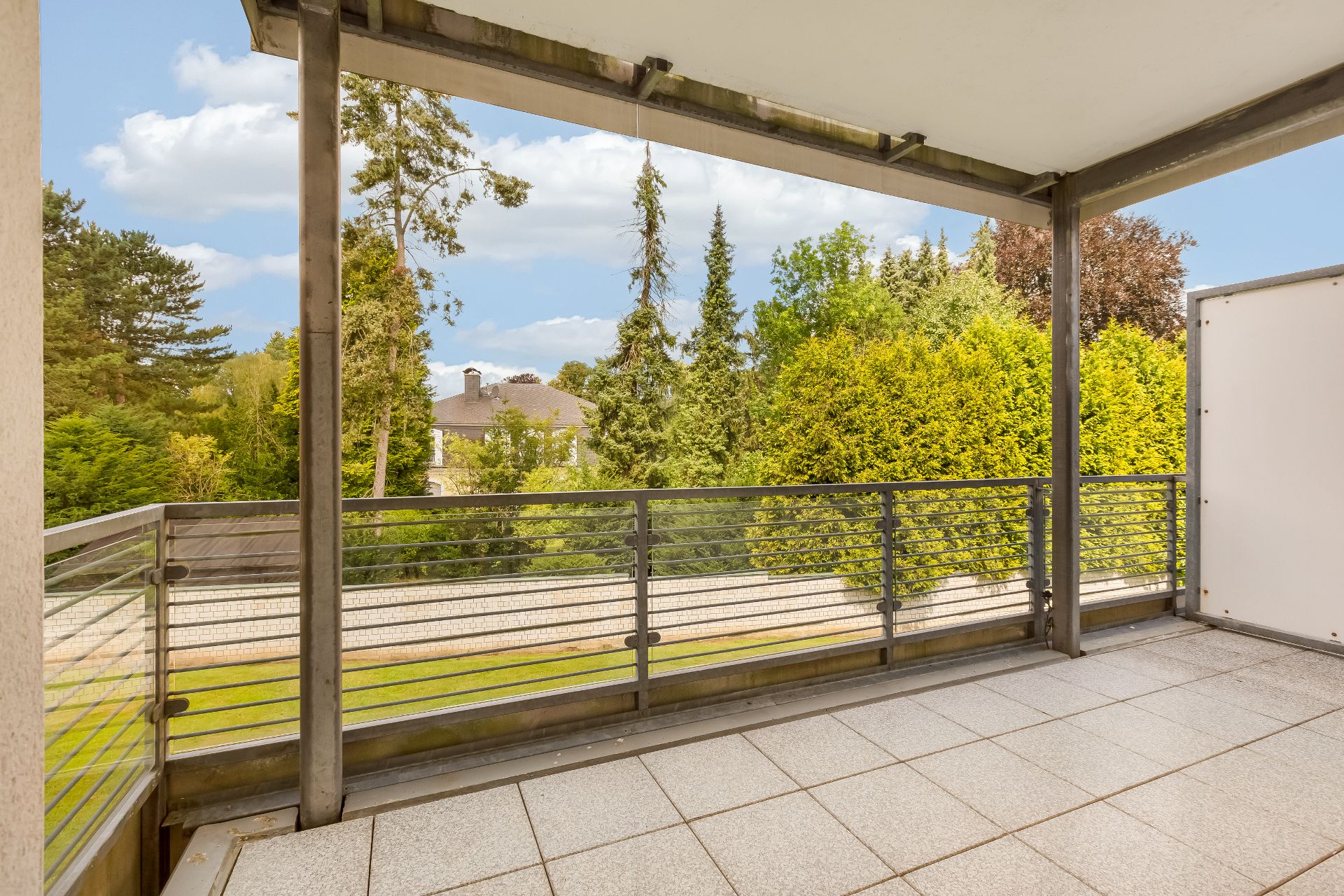 Überdachter Balkon mit Geländer und Blick auf grüne Bäume und einen Garten.
