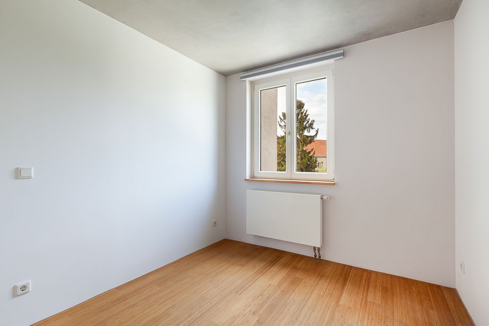 Leeres Zimmer mit weißer Wand, Holzboden, Fenster und Heizkörper.
