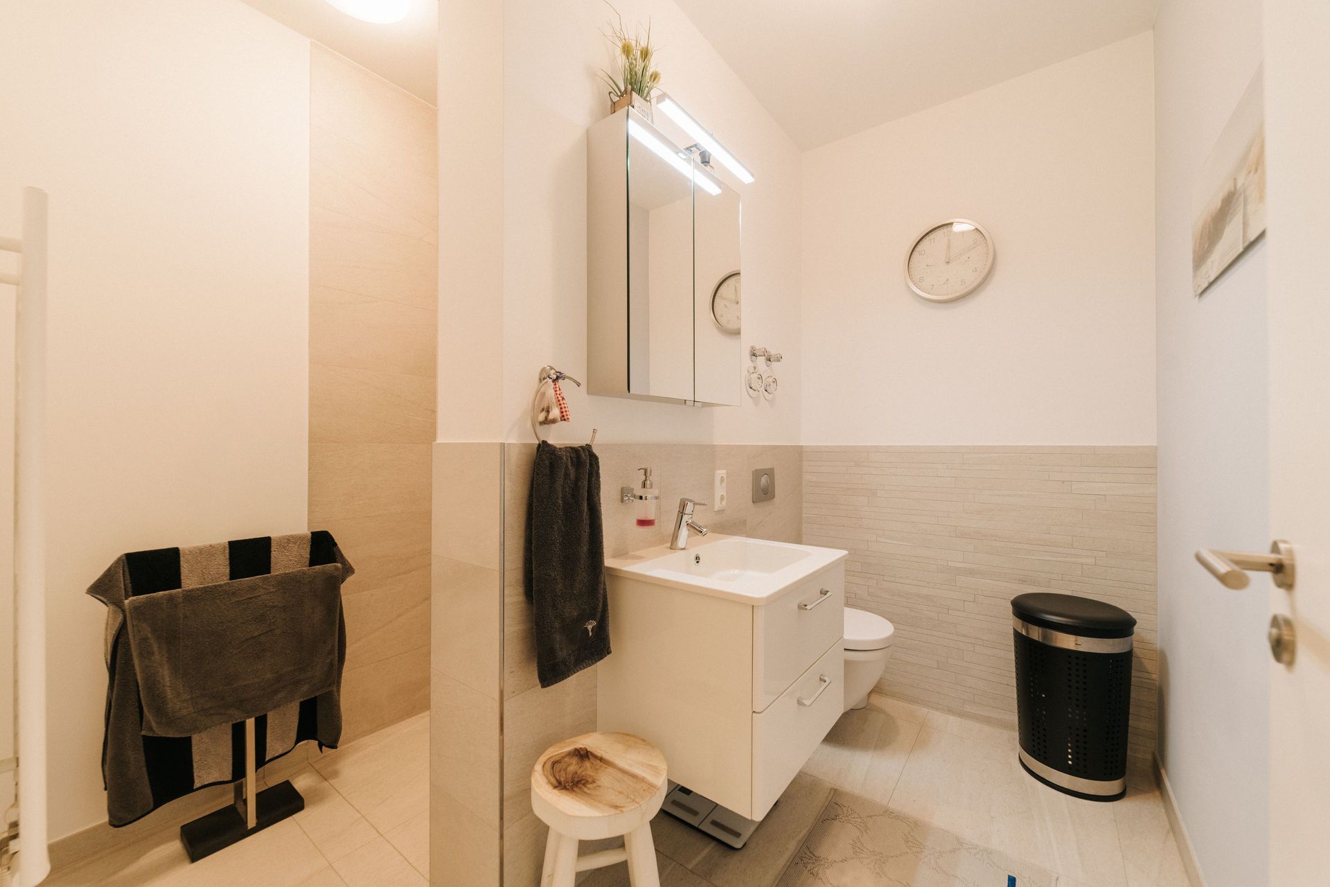 Modernes Badezimmer mit Waschbecken, Spiegel, Hocker, Handtuchständer, Uhr und Mülleimer.