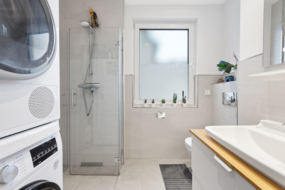 Modernes Badezimmer mit Dusche, Waschmaschine, Trockner, Waschbecken und Fenster mit Pflanzen.
