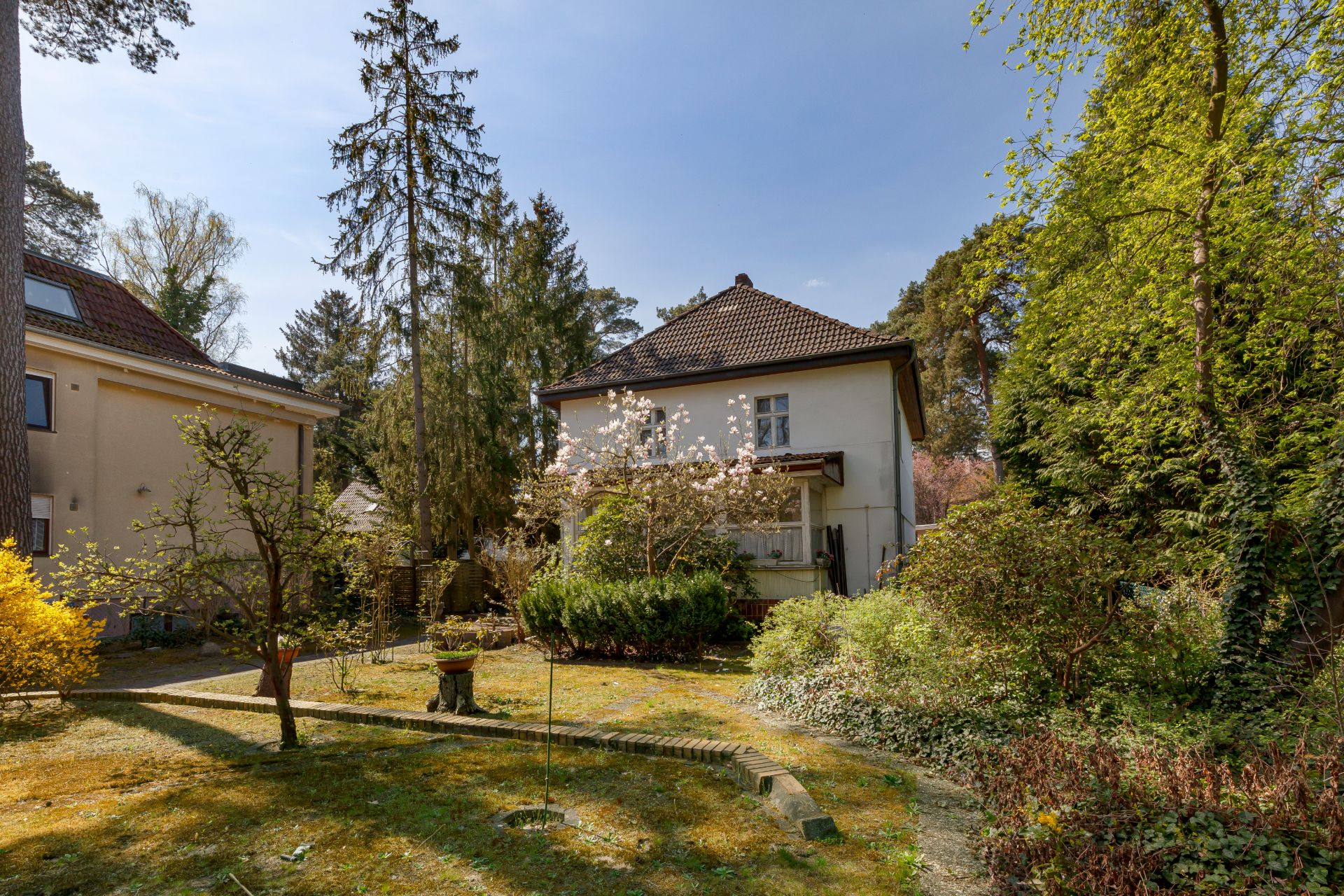 Zweistöckiges Wohnhaus mit Garten, umgeben von Bäumen und Sträuchern an einem sonnigen Tag.
