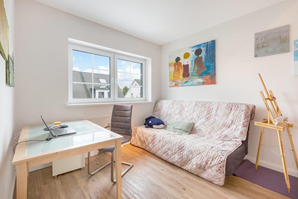 Helles Zimmer mit Schreibtisch, Stuhl, Sofa, Staffelei und Bildern an der Wand.