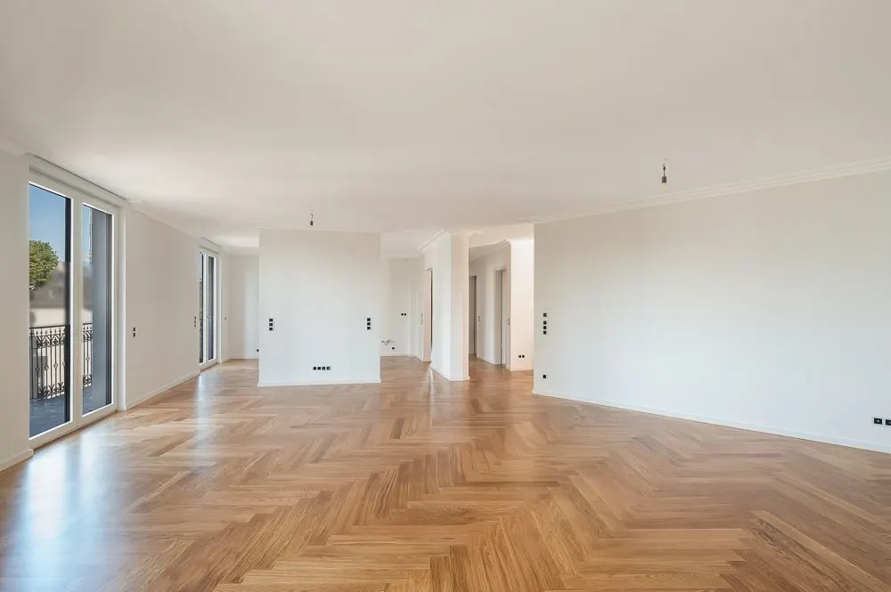Großer, leerer Raum mit Parkettboden und bodentiefen Fenstern in einer Wohnung.