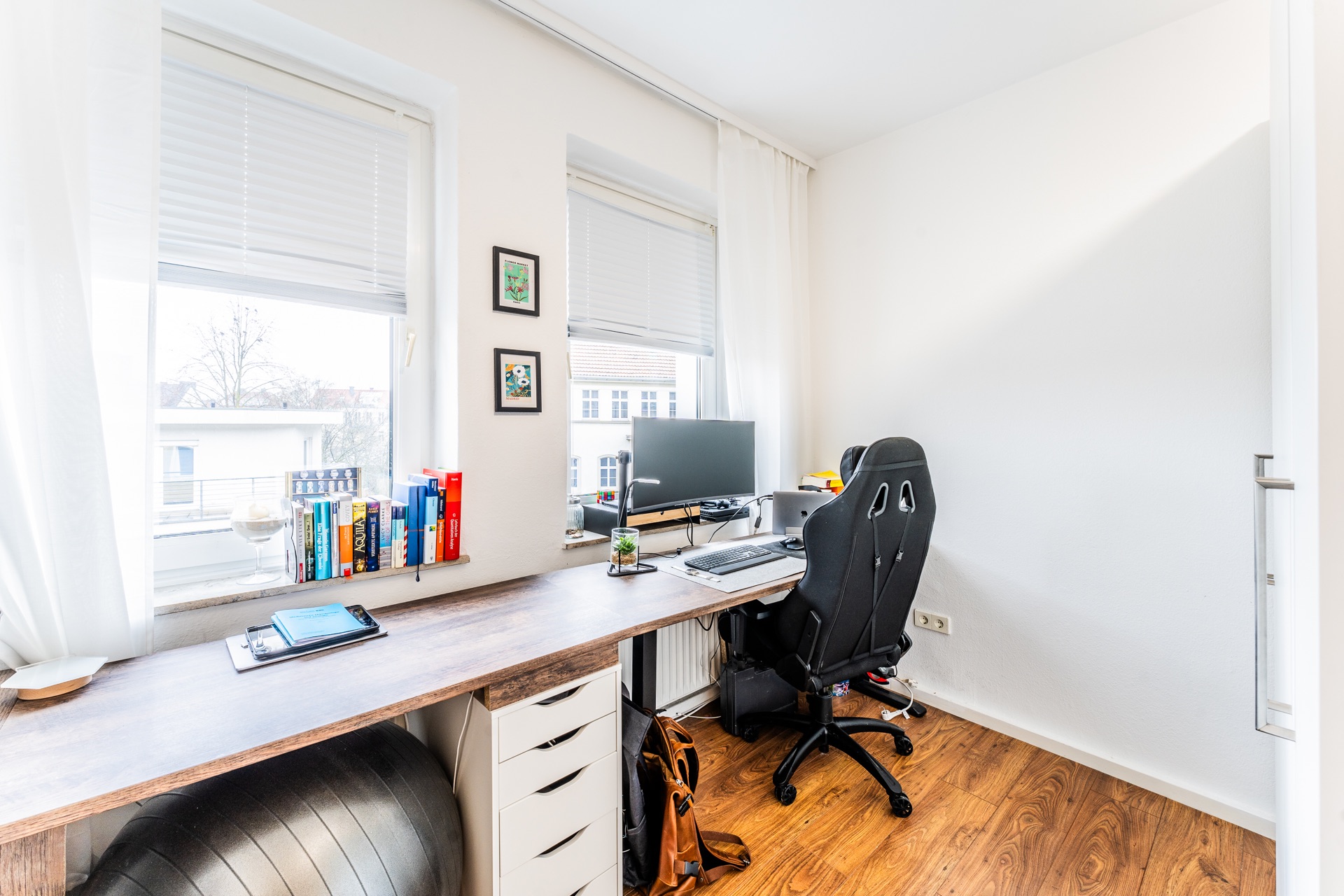 Modernes Arbeitszimmer mit Schreibtisch, Monitor, Bürostuhl und Bücherregal am Fenster.