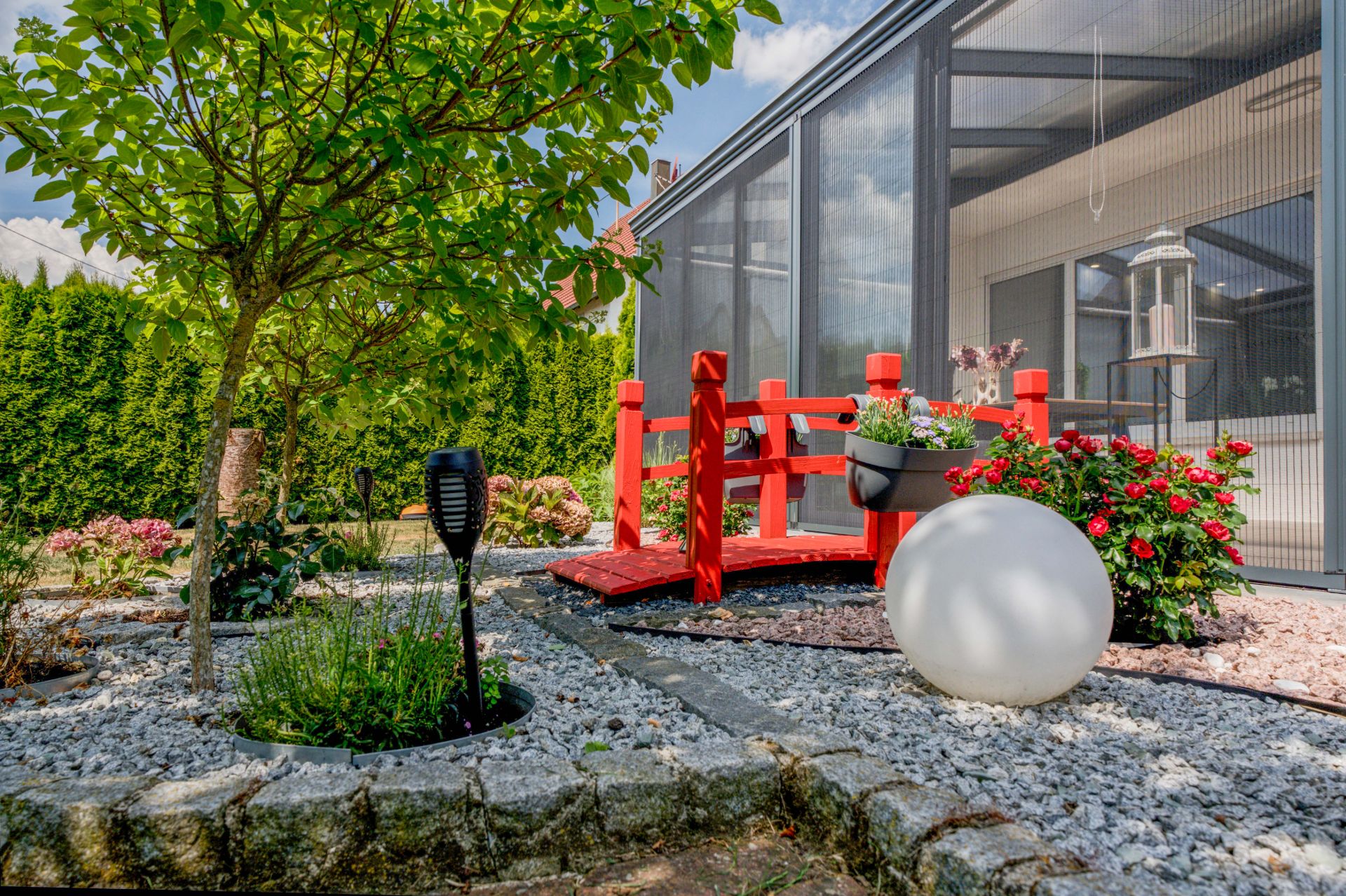 Garten mit kleinem Baum, roter Brücke, Blumen und großer runder Lampe vor einem Wintergarten.