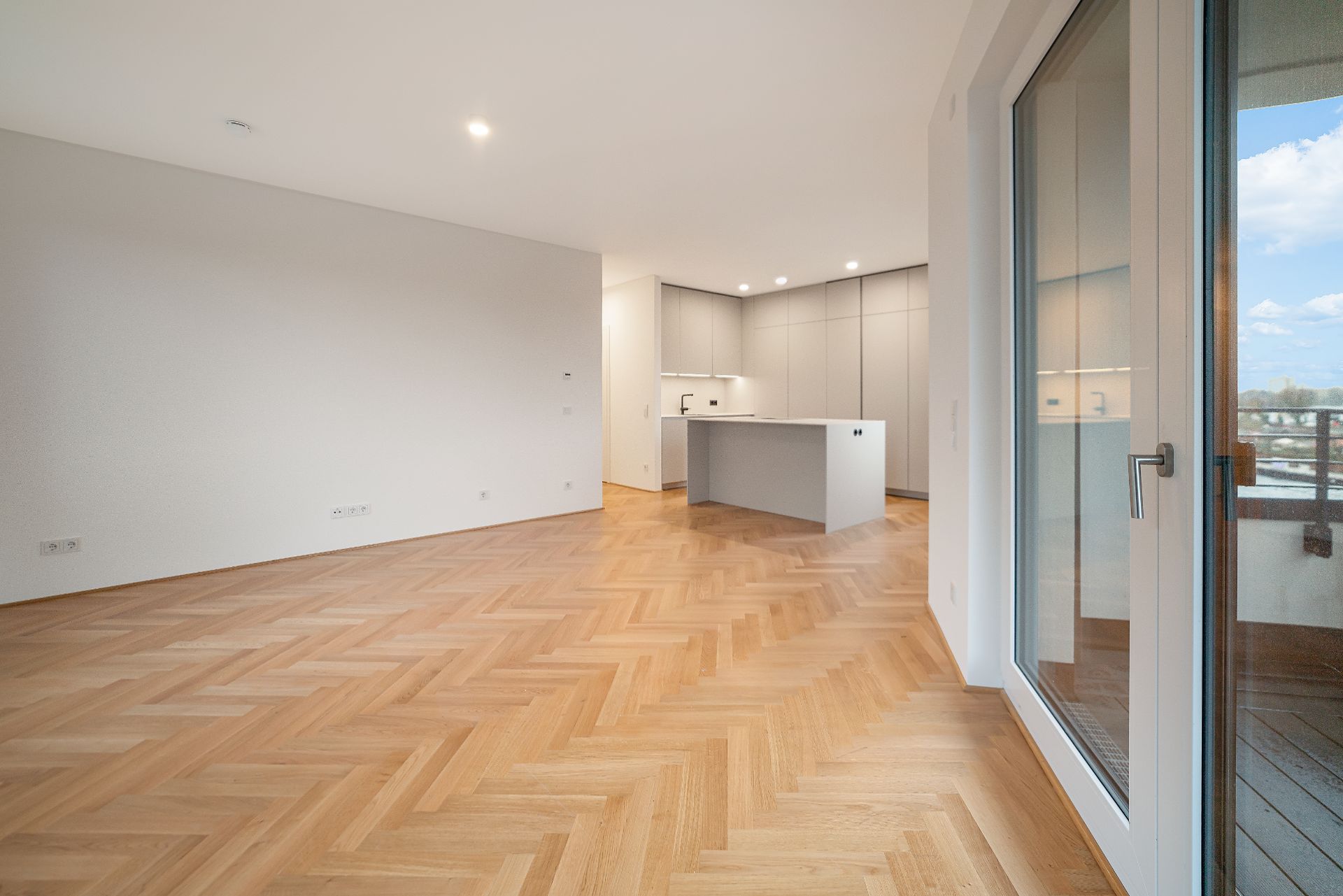 Heller Raum mit Parkettboden, offener Küche und großen Fenstern zu einem Balkon.