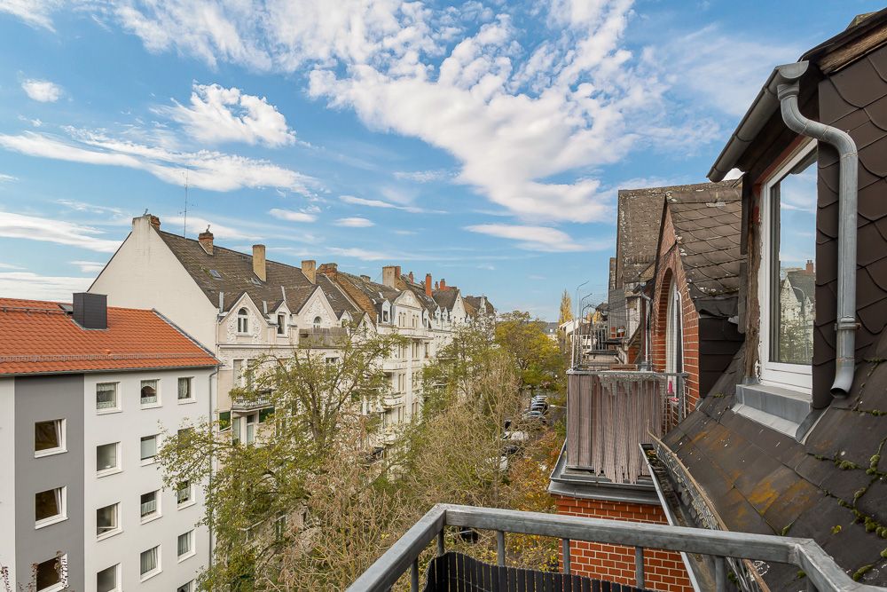Blick von einem Balkon auf eine Baumallee und Wohnhäuser unter blauem Himmel mit Wolken.