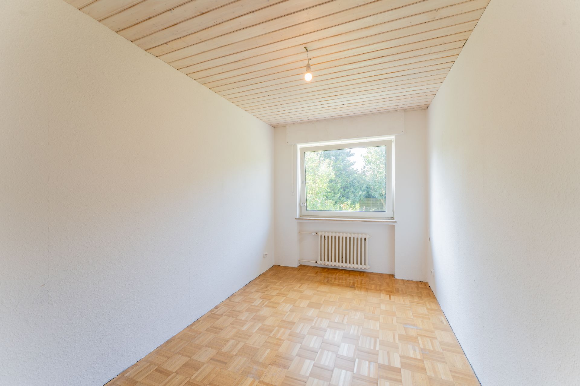 Heller, leerer Raum mit Holzdecke, Parkettboden und Fenster mit Blick ins Grüne.