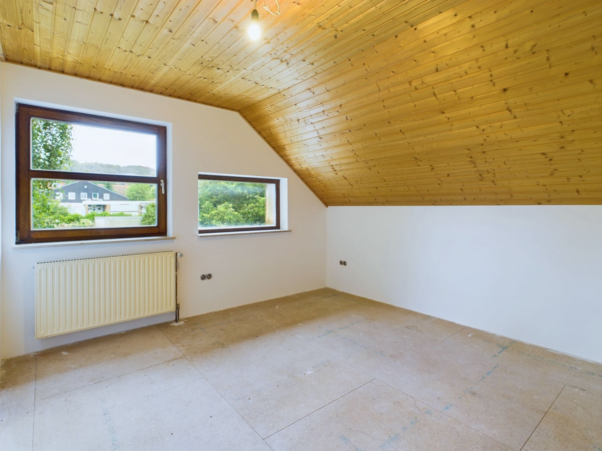 Leerer Dachzimmerraum mit Holzdecke, weißen Wänden und zwei Fenstern.