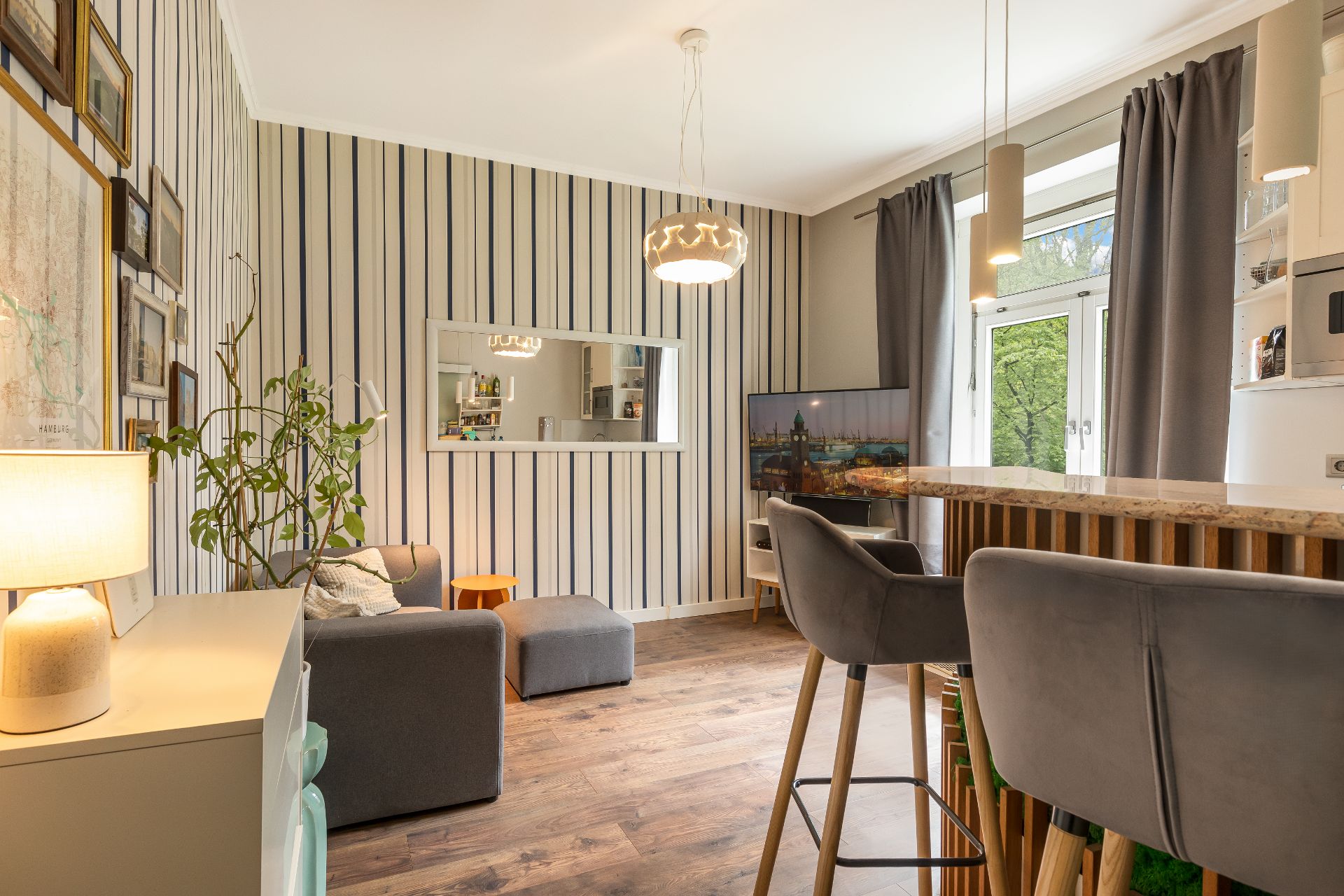 Helles Wohnzimmer mit gestreifter Tapete, Sessel, Barhockern, TV und großem Fenster mit Vorhängen.