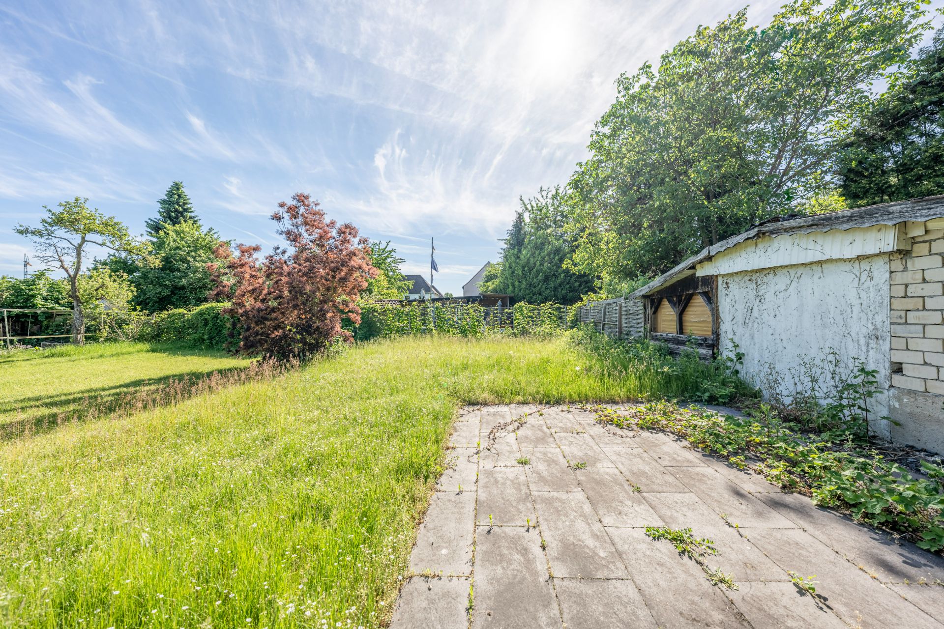 Garten mit verwildertem Rasen, Bäumen, Terrasse und einem Schuppen rechts im Bild.