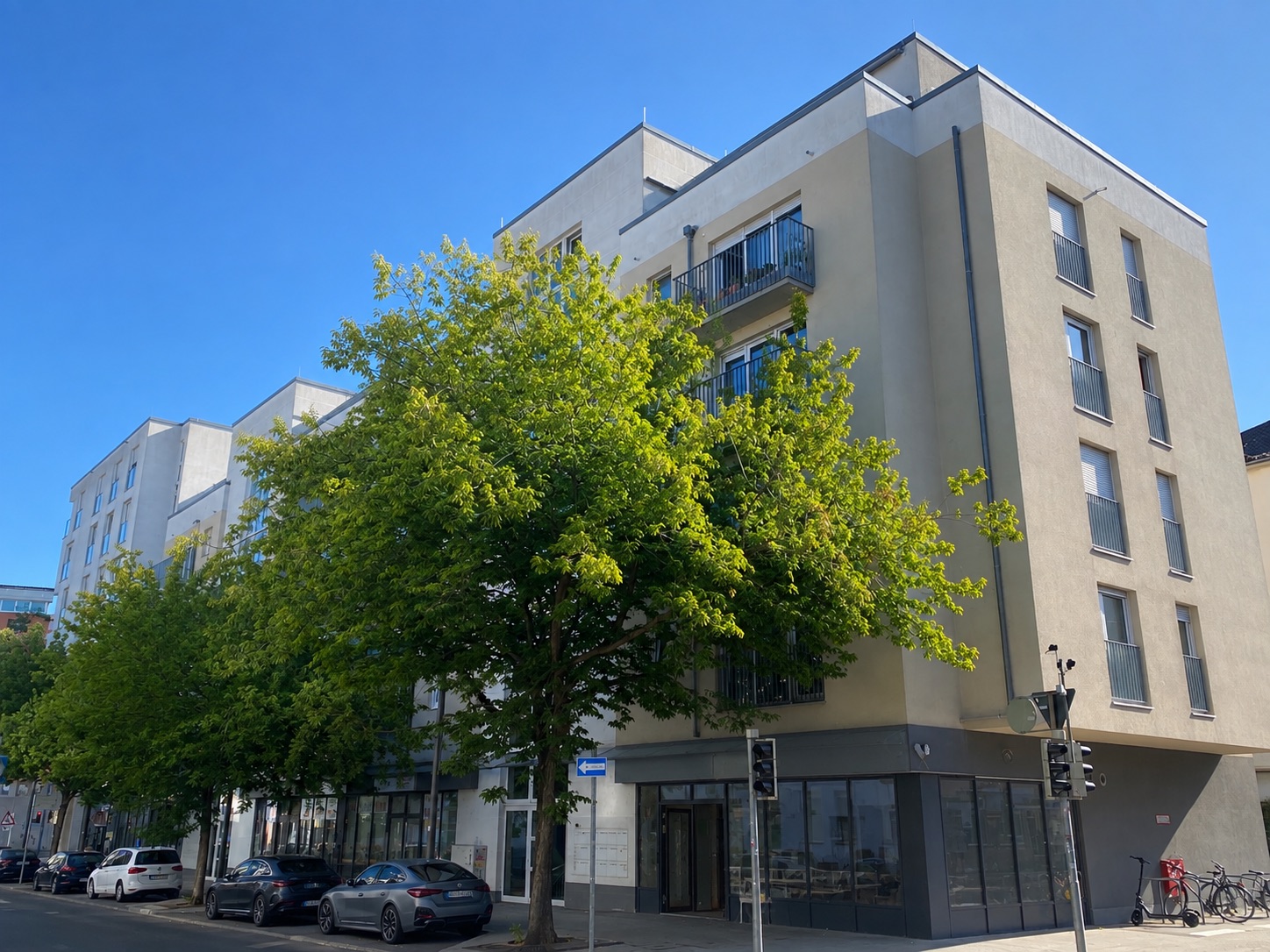 Modernes Wohnhaus an einer Straßenecke mit Bäumen und geparkten Autos.