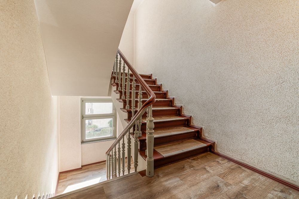 Holztreppe mit Handlauf in einem hellen Treppenhaus, Fenster im Hintergrund.
