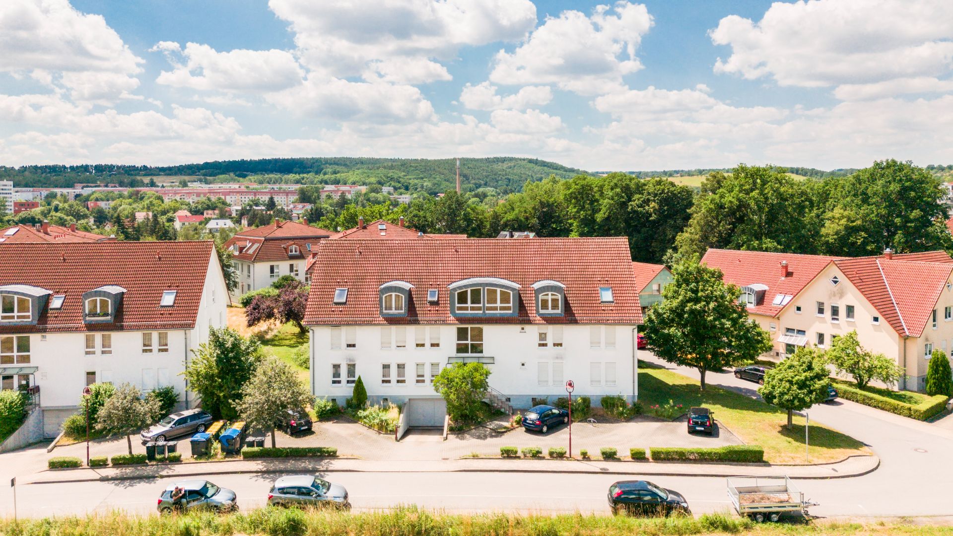 Mehrfamilienhäuser mit roten Dächern, Bäumen und geparkten Autos bei sonnigem Wetter.