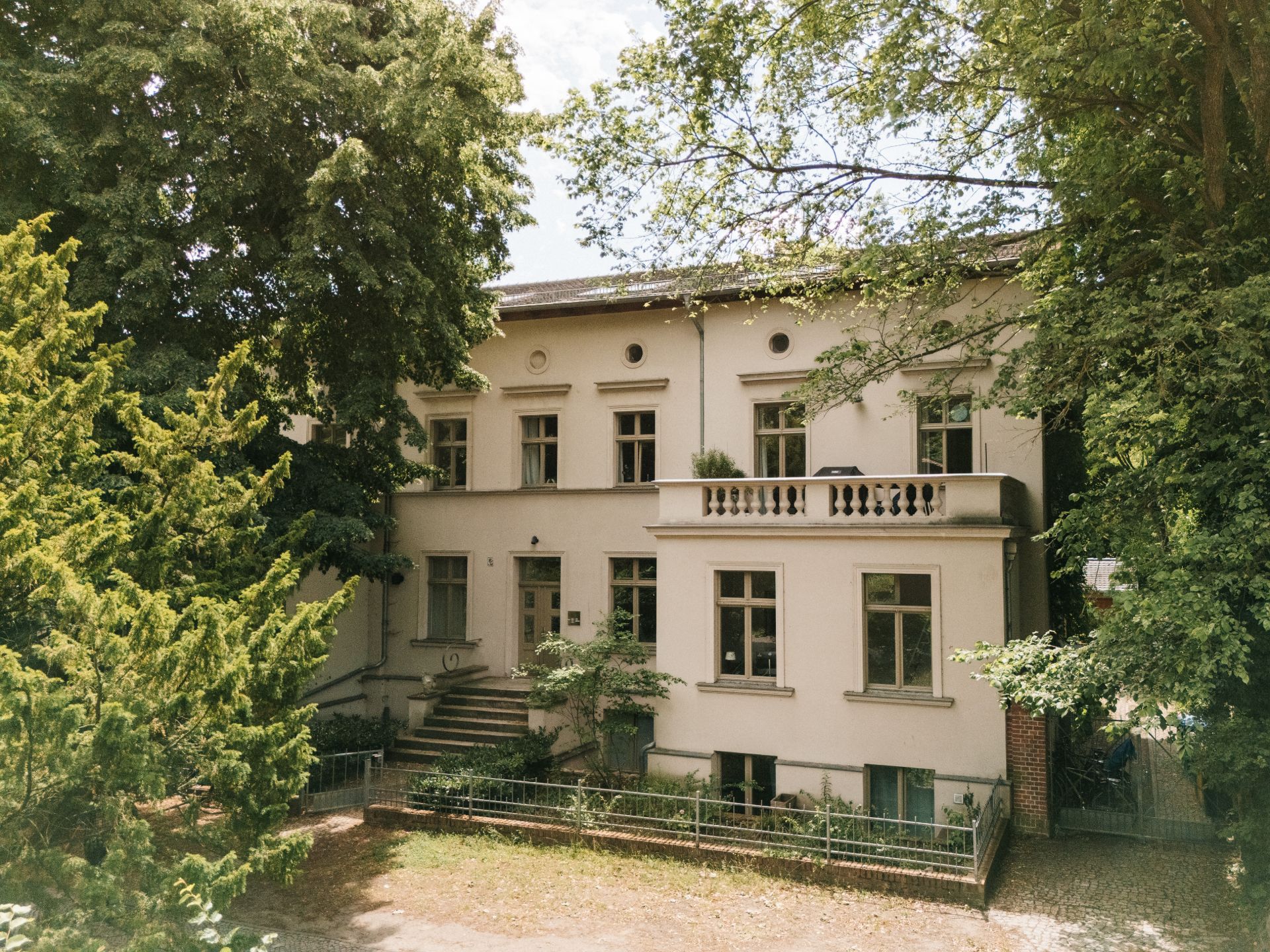 Zweistöckiges Wohnhaus mit mehreren Fenstern, umgeben von Bäumen und Grünflächen.