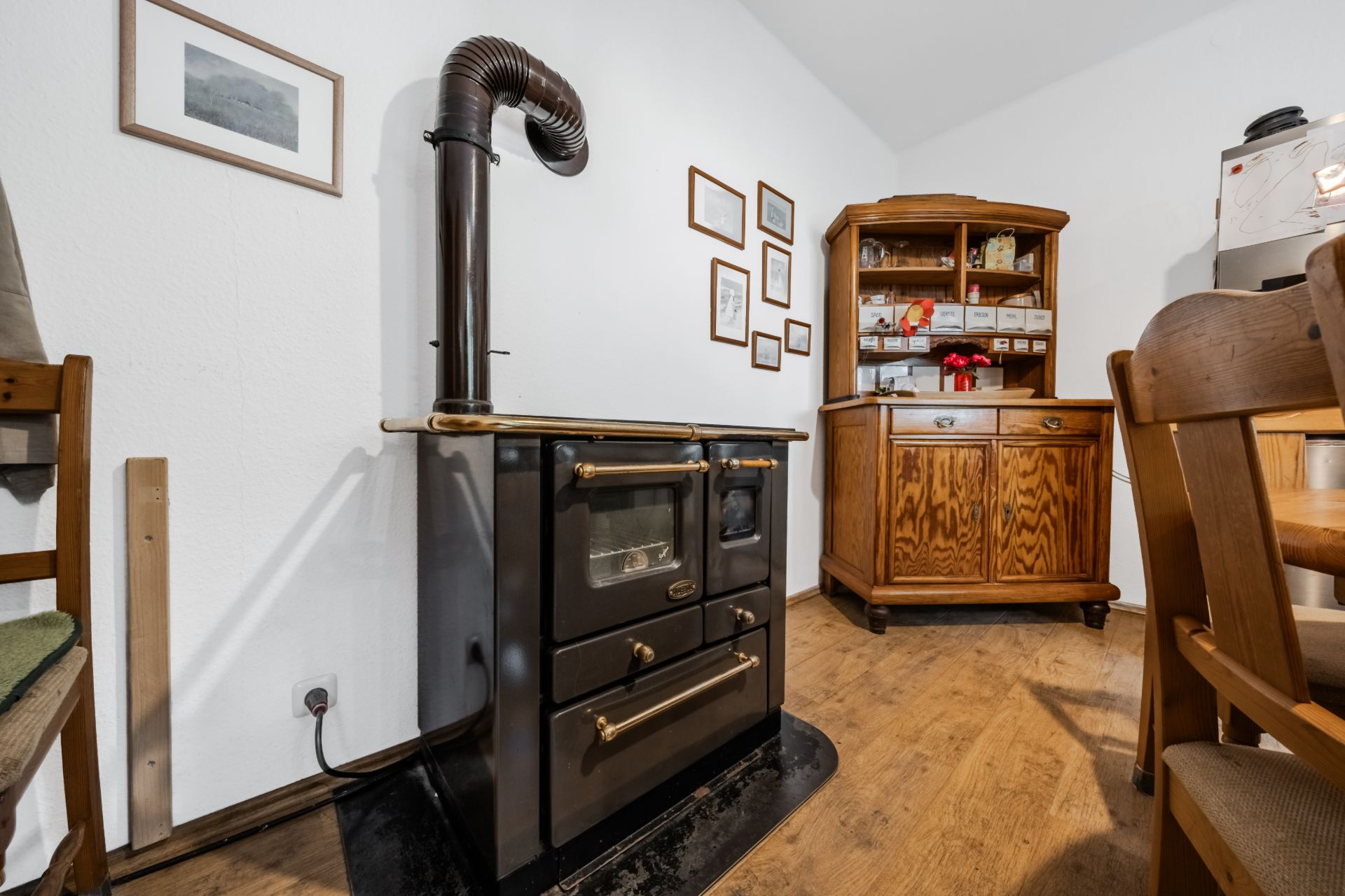 Ein Holzofen steht in einer gemütlichen Wohnküche mit Holzmöbeln und Bilderrahmen an der Wand.