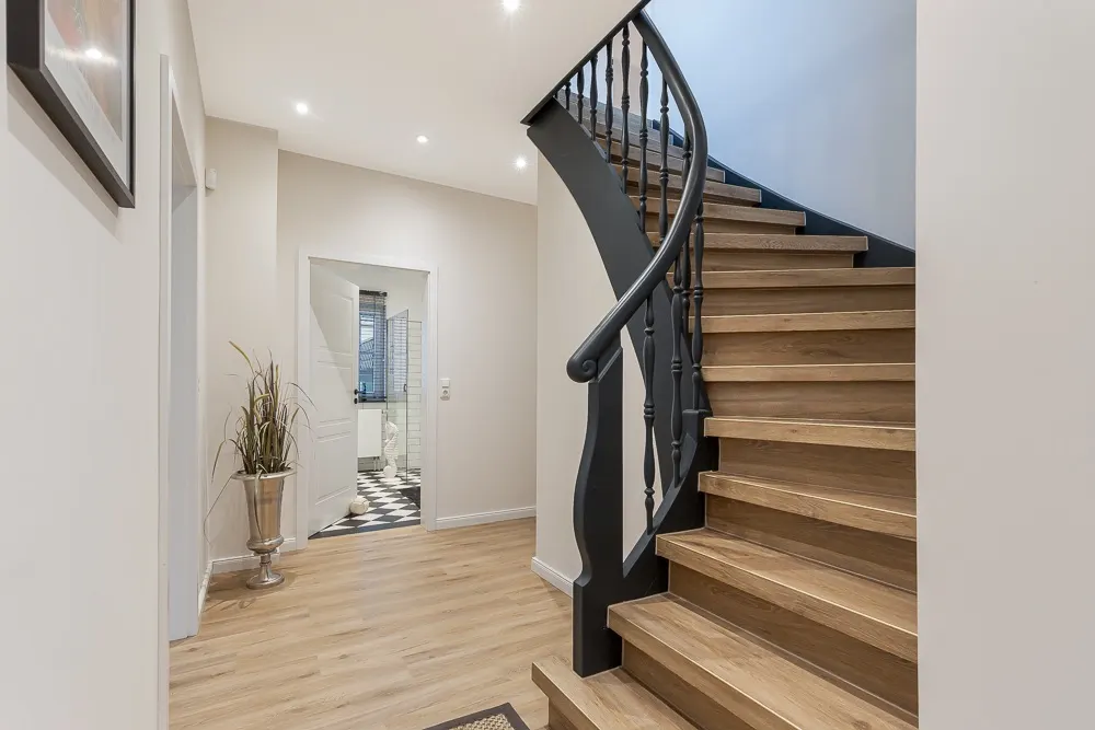 Heller Flur mit Holztreppe, Pflanze in Vase und Blick ins Badezimmer mit Fliesenboden.