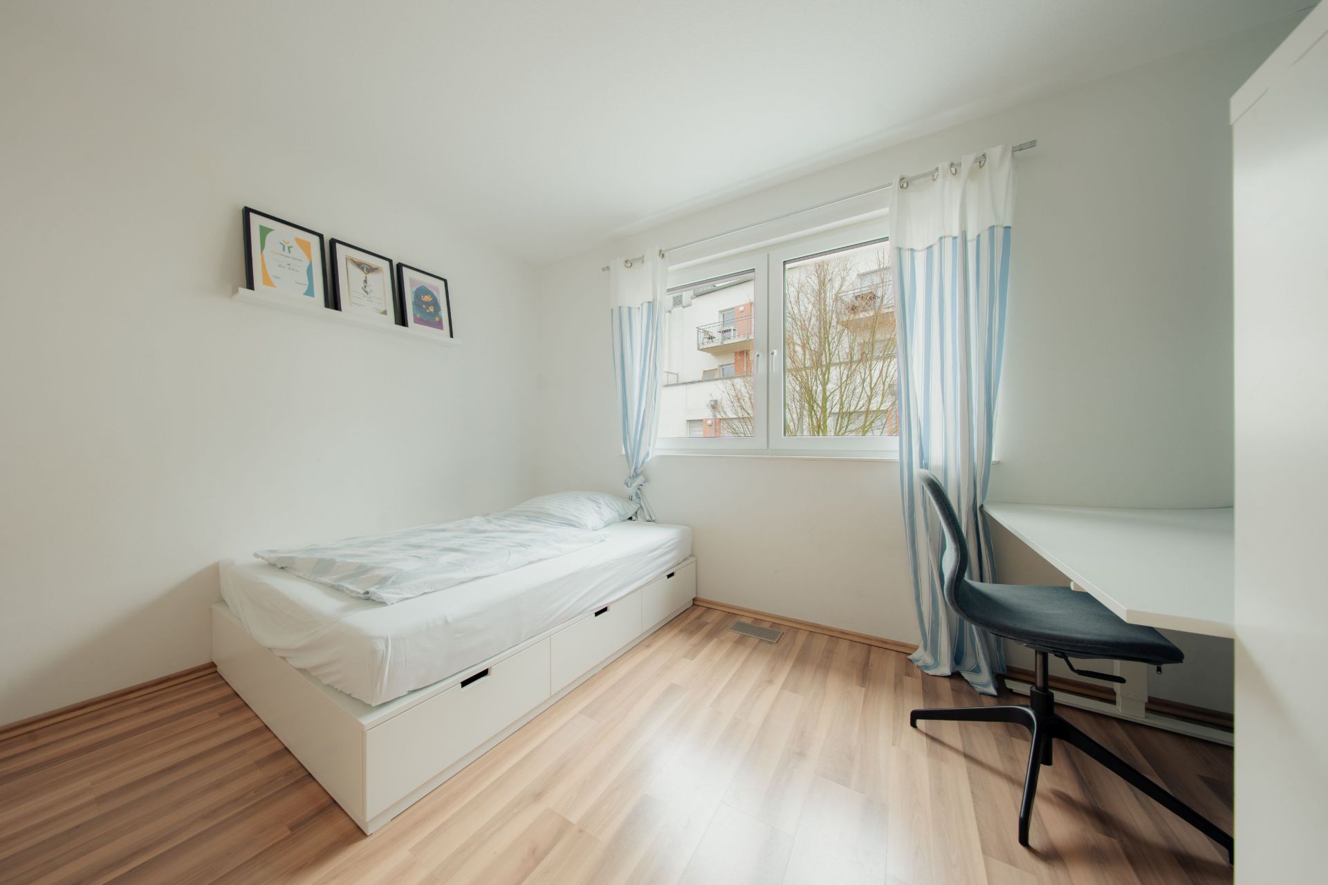 Helles Schlafzimmer mit Einzelbett, Schreibtisch, Stuhl und Fenster mit Vorhängen.