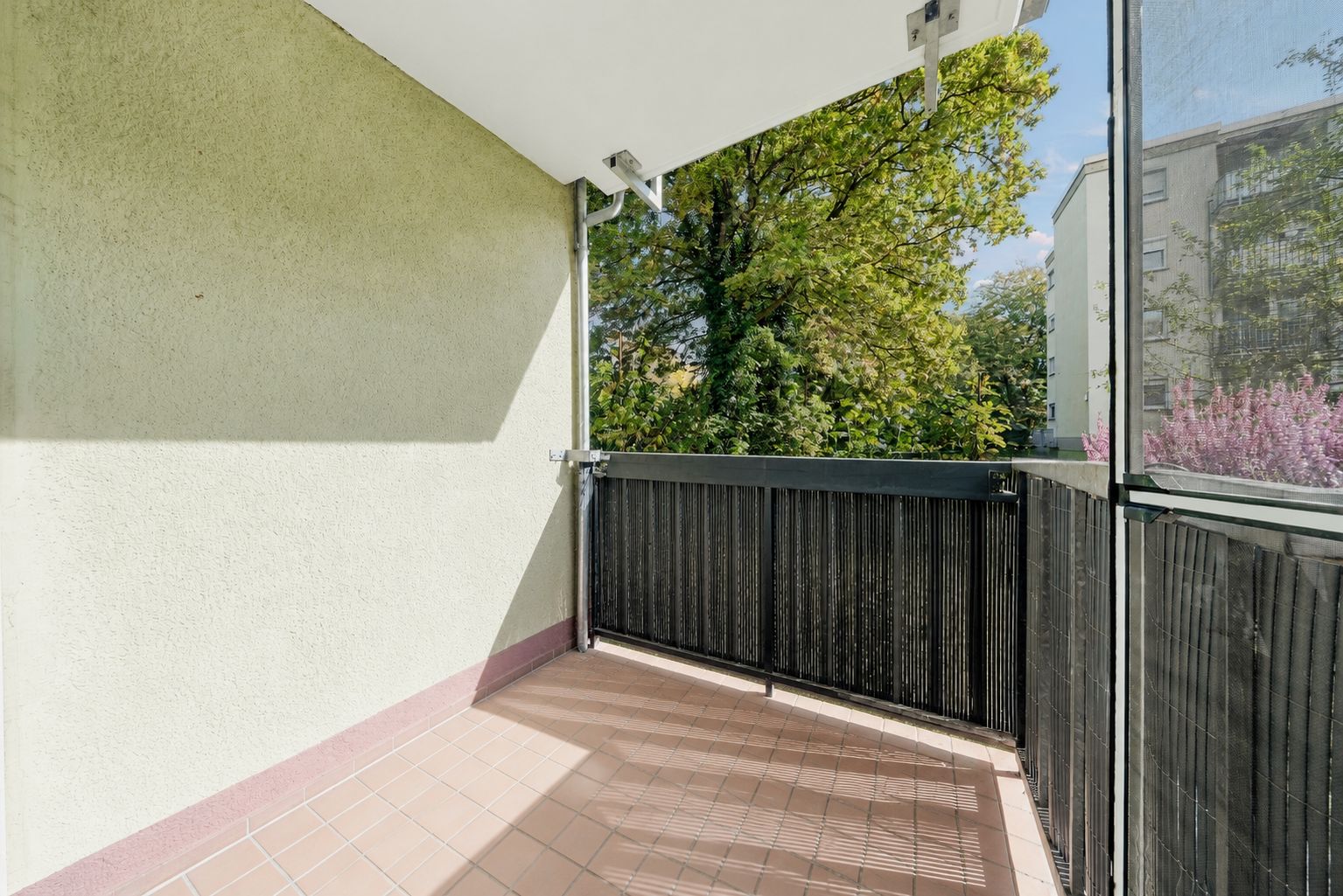 Überdachter Balkon mit Fliesenboden, schwarzem Geländer und Blick auf Bäume.