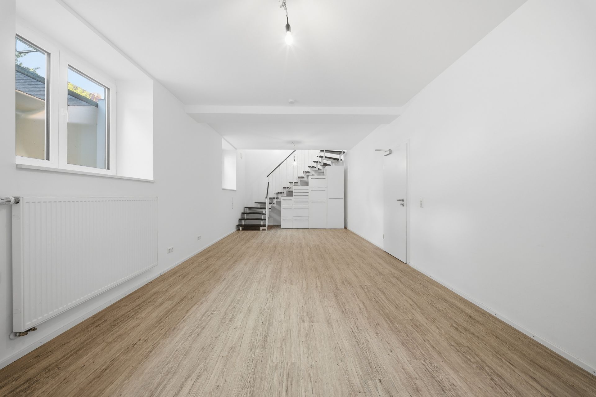 Heller Raum mit Holzboden, weißen Wänden, Fenstern und einer modernen Treppe im Hintergrund.