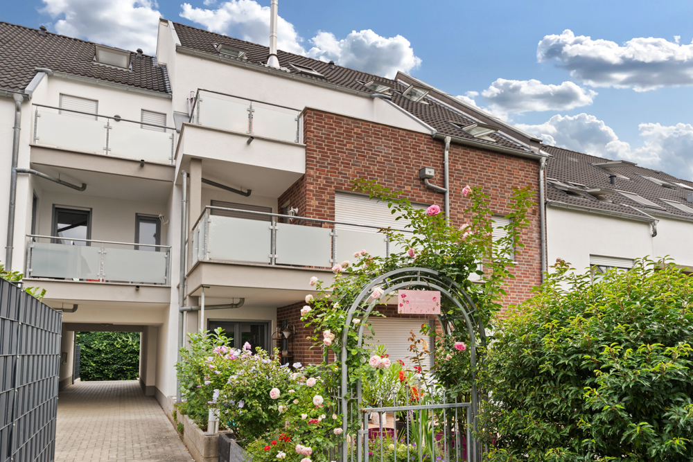 Modernes Wohngebäude mit Balkon, Backsteinfassade und blühendem Vorgarten mit Rosenbogen.
