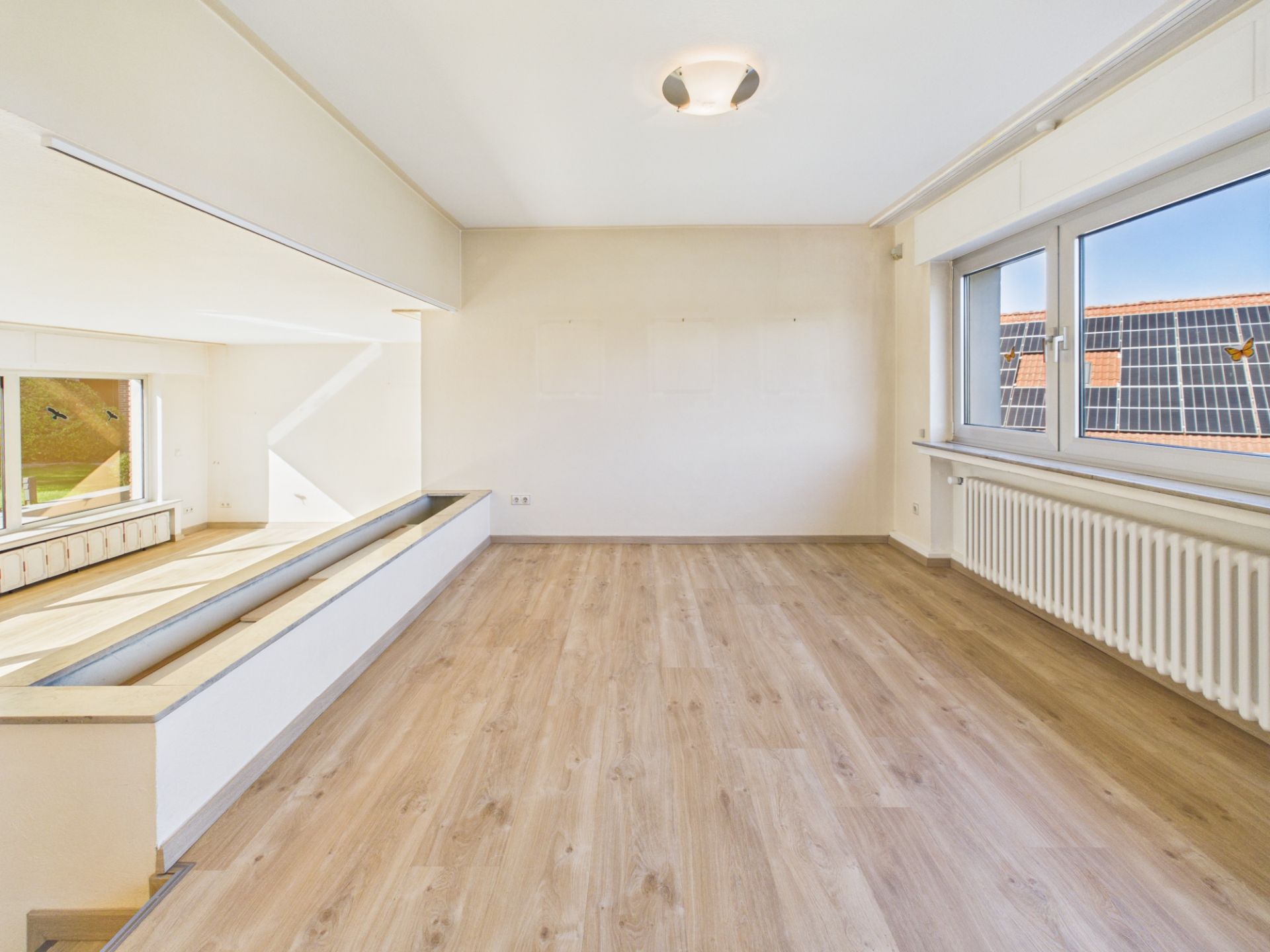 Heller, leerer Raum mit Parkettboden, großen Fenstern und Heizkörper unter dem Fenster.