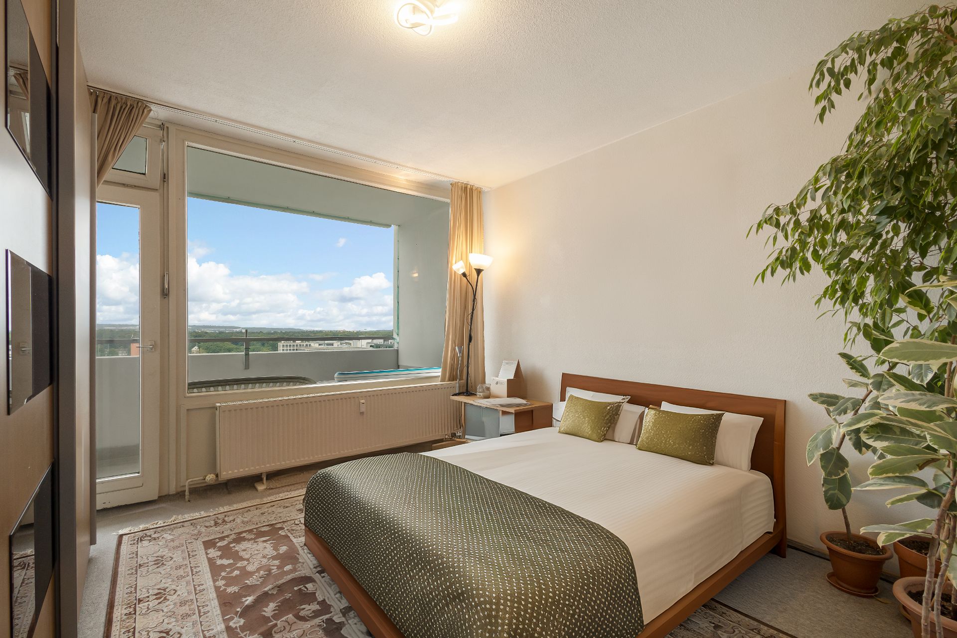 Helles Schlafzimmer mit Doppelbett, großem Fenster, Teppich und Zimmerpflanze.