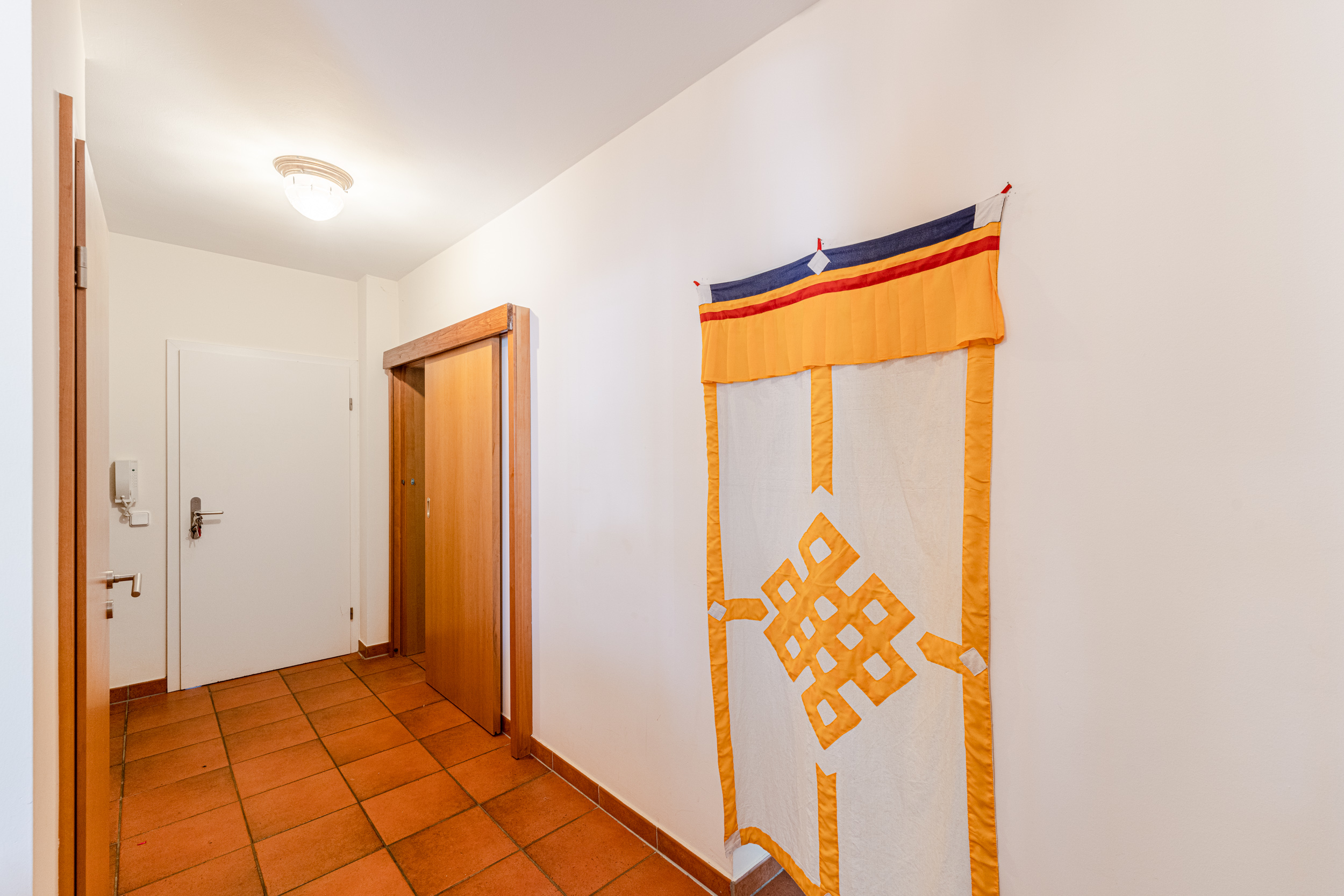 Flur mit braunen Fliesen, weißer Tür, Holzschrank und bunter Wanddeko.