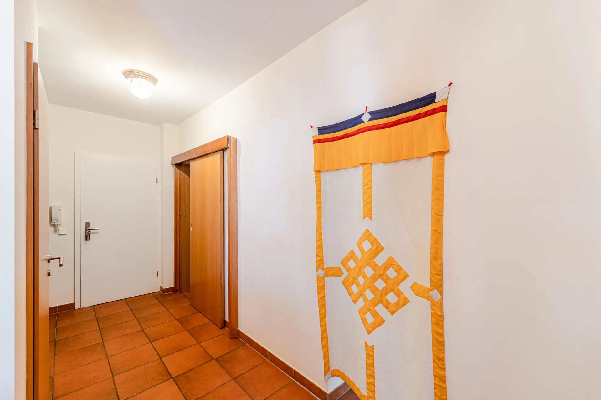 Flur mit braunen Fliesen, weißer Tür, Holzschrank und bunter Wanddeko.