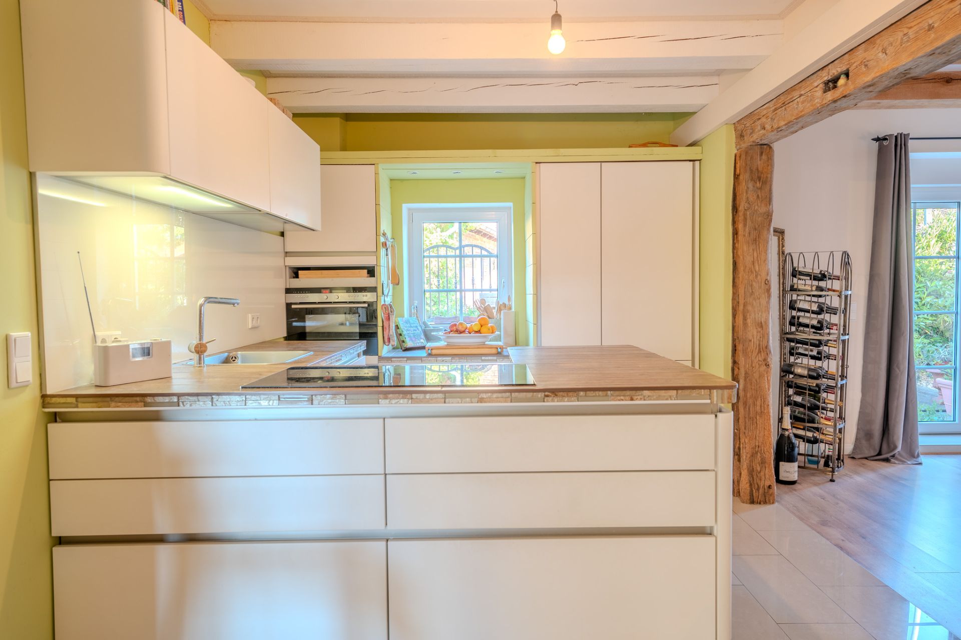 Moderne Küche mit weißer Einrichtung, Insel, großem Fenster und Blick auf einen Weinständer im Wohnbereich.
