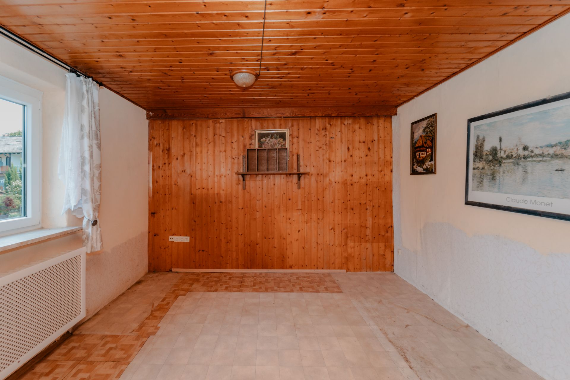 Leerer Raum mit Holzdecke, Holzvertäfelung, zwei Bildern und Fenster mit weißen Gardinen.