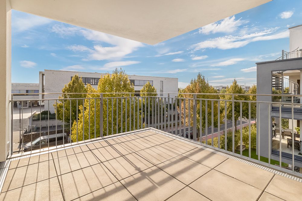 Balkon mit Fliesenboden und Metallgeländer, Blick auf Bäume und moderne Wohngebäude.