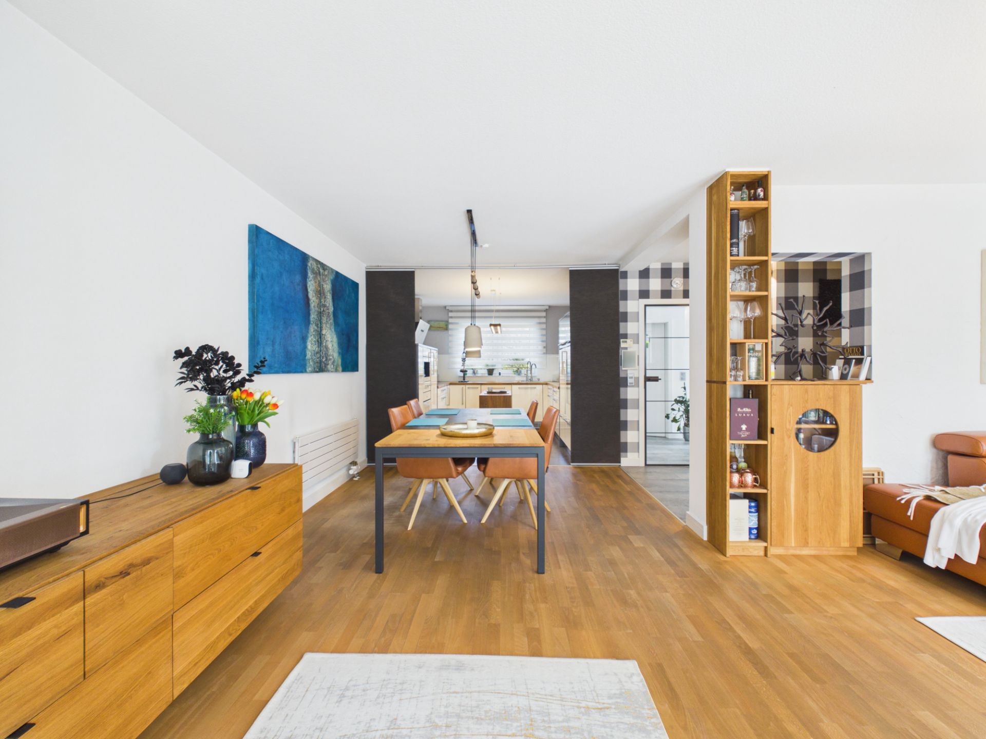 Helles Wohnzimmer mit Holzboden, Esstisch, Sideboard und Regal, Blick zur offenen Küche.